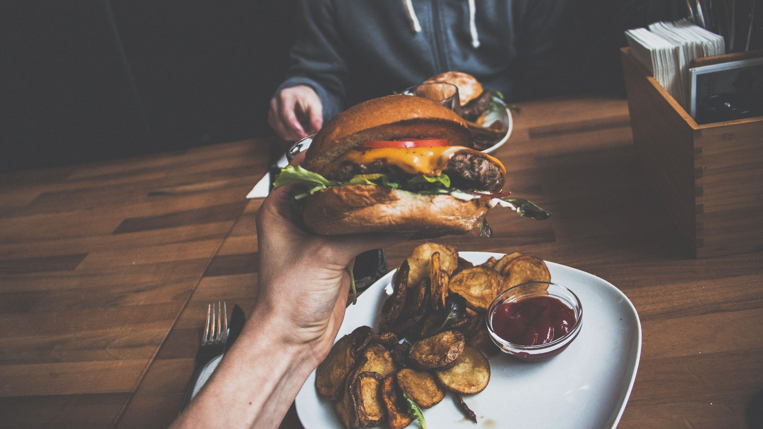 A close-up of a hand holding a burger filled with lettuce, tomato, and cheese above a white plate with potato wedges and a small container of ketchup. Another person's hands are partially visible in the background, also holding a burger. The scene is set on a wooden table.