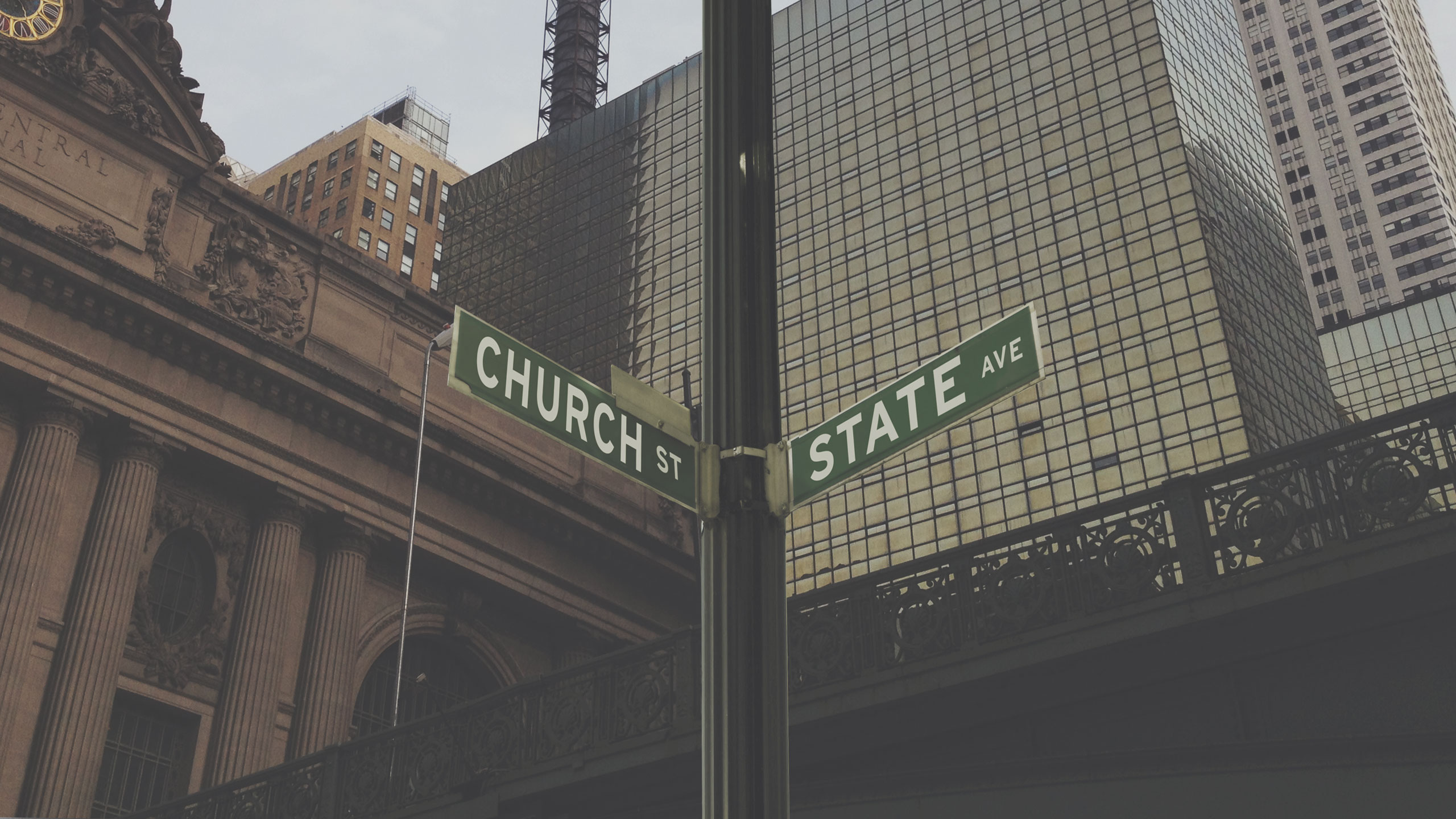 A street sign positioned at the corner of Church Street and State Avenue in an urban setting with tall buildings, including one with a classical architectural style, and reflective glass windows in the background.