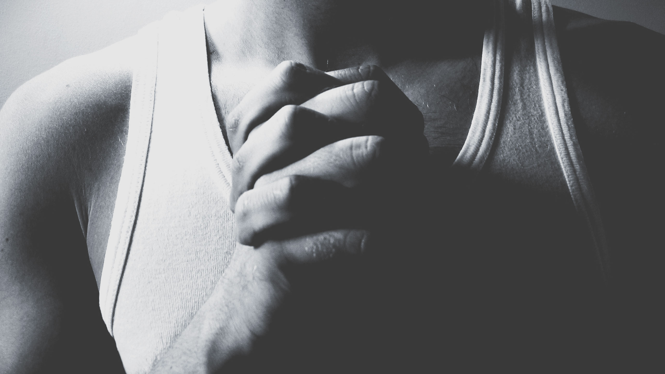 A black-and-white close-up image of a person wearing a sleeveless top, with their hands clasped together in front of their chest. Only the lower part of their face, neck, and upper chest are visible. The image conveys a contemplative or prayerful mood.