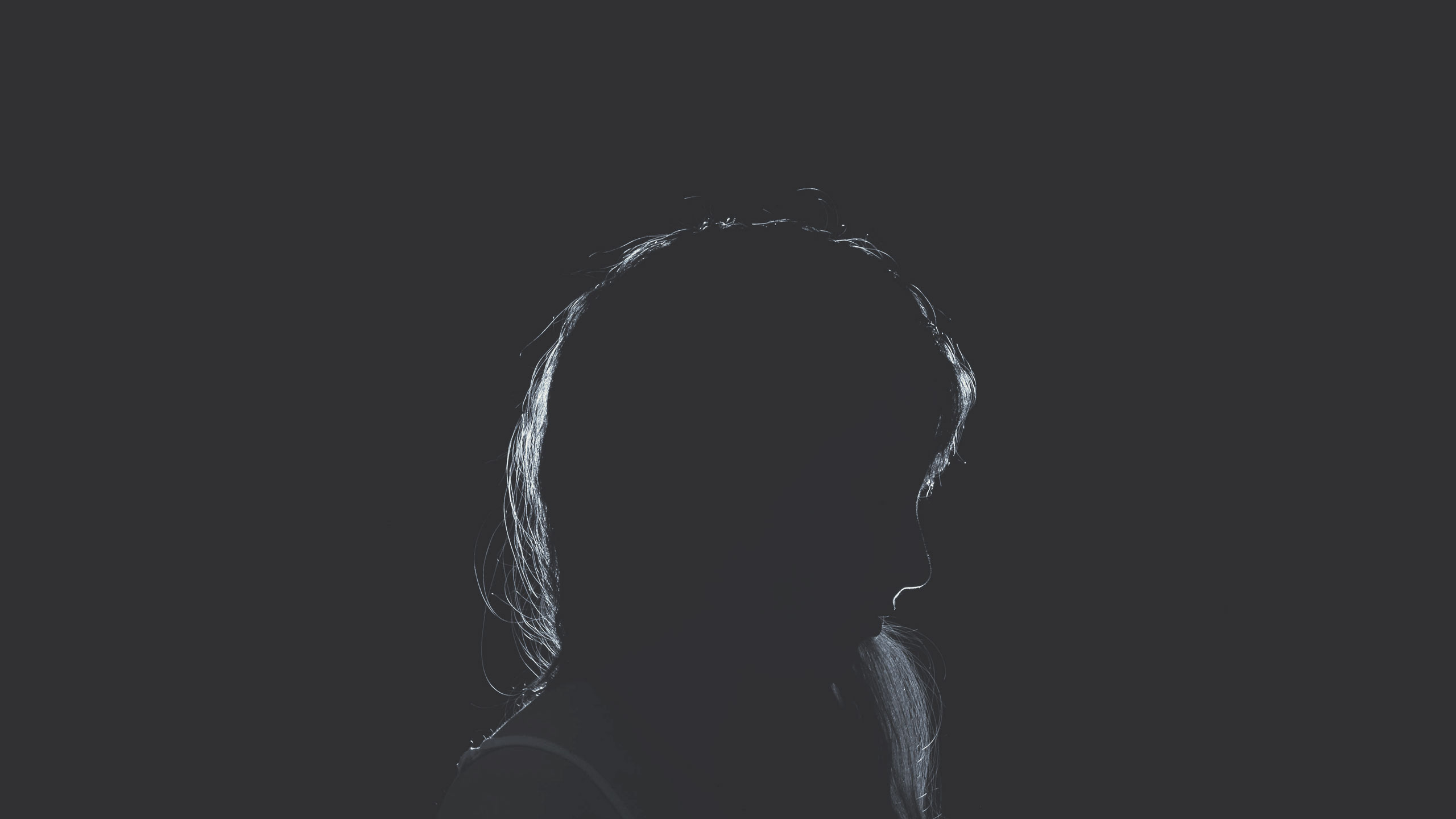 A silhouette of a person with long hair captured against a dark background. The lighting highlights the outline of their hair and head, creating a dramatic, mysterious effect. The figure is turned slightly to the side, looking downward.