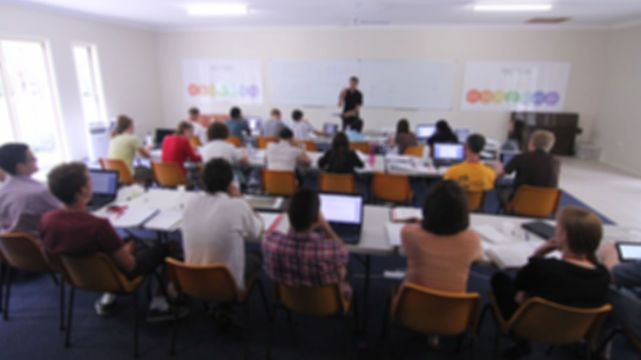 A classroom setting with students seated at tables facing a whiteboard. The students are using laptops, notebooks, and other learning materials. A teacher stands at the front of the room, engaging with the students. The image is blurry.