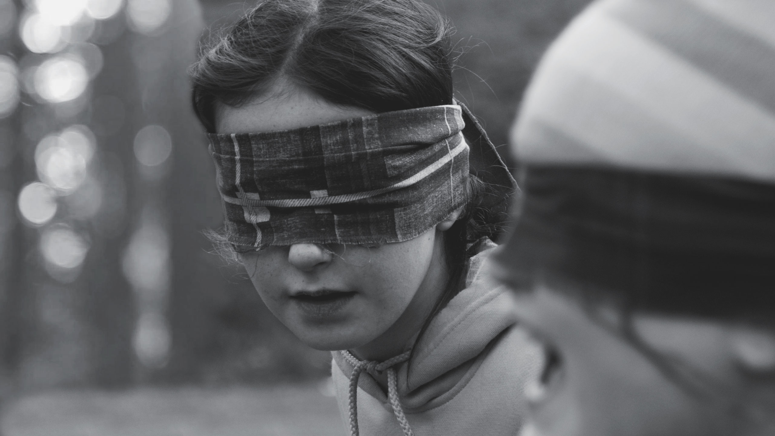 Black and white photo of two people. The person in the foreground is wearing a plaid blindfold and a hooded sweatshirt, concentrating intently. The second person, to the right and slightly out of focus, is also blindfolded. The background is blurry, suggesting an outdoor setting.