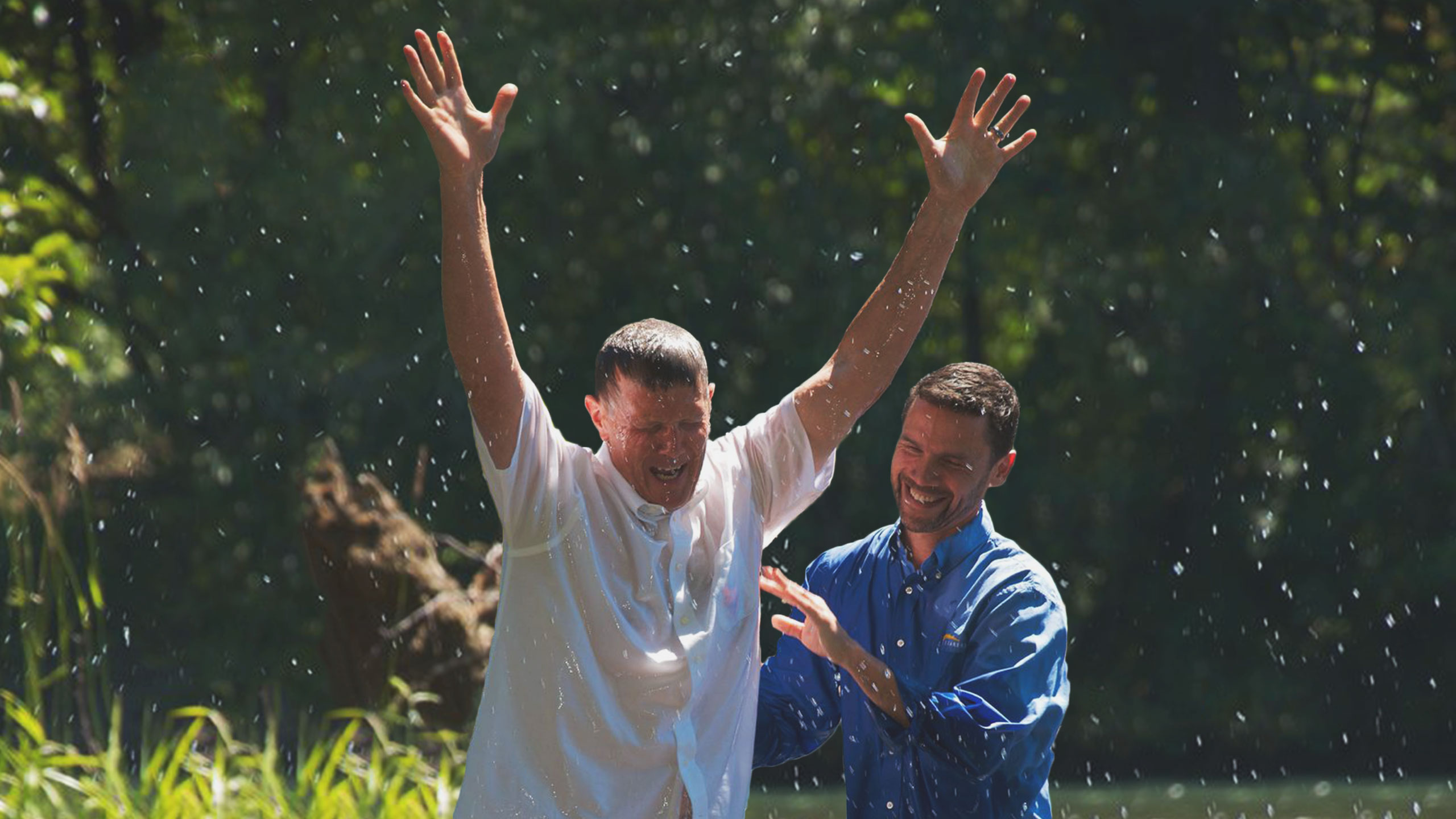 Two men are in a river, surrounded by lush greenery. One man, in a white shirt, has his arms raised joyfully as water droplets fly around. The other man, in a blue shirt, smiles and touches the first man's arm. The scene suggests a celebratory moment, likely a baptism.