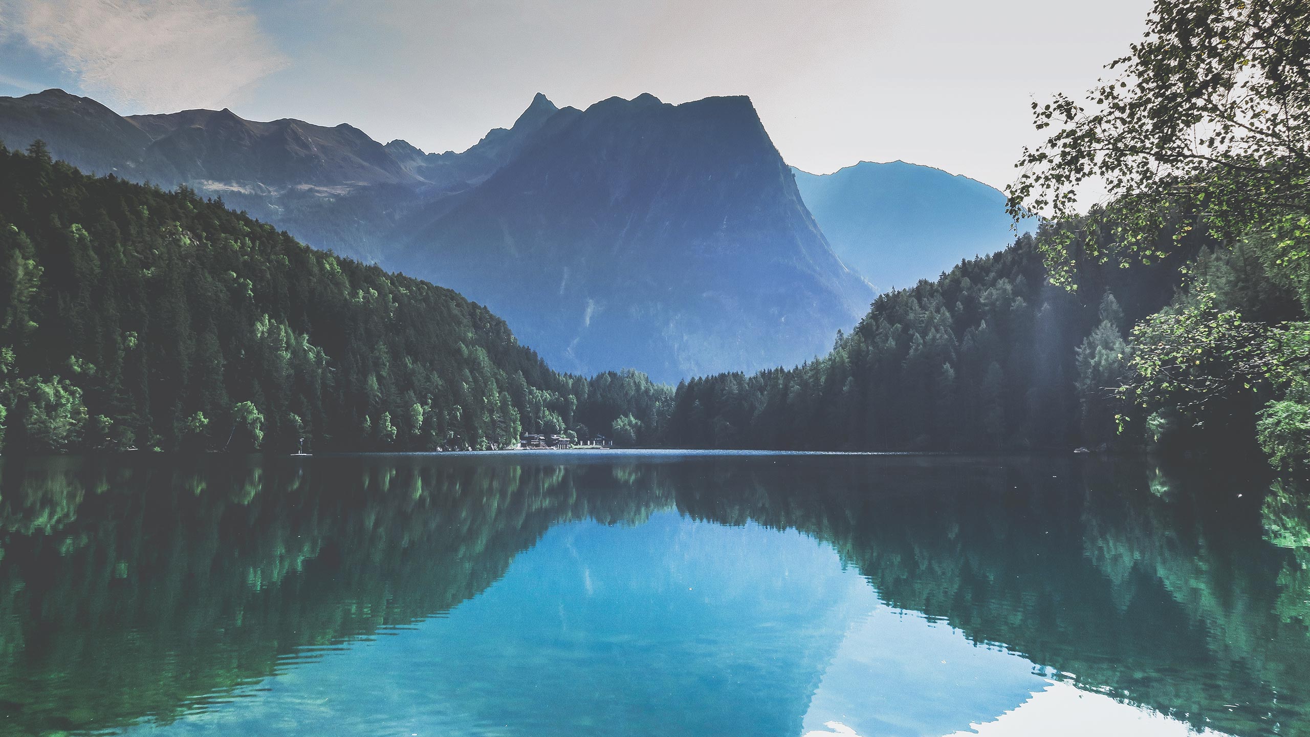 A serene mountain lake is surrounded by dense evergreen forests. Mountains in the background are shrouded in mist. The lake's glassy surface reflects the trees and sky, creating a mirror-like effect. Soft sunlight filters through, illuminating the tranquil scene.