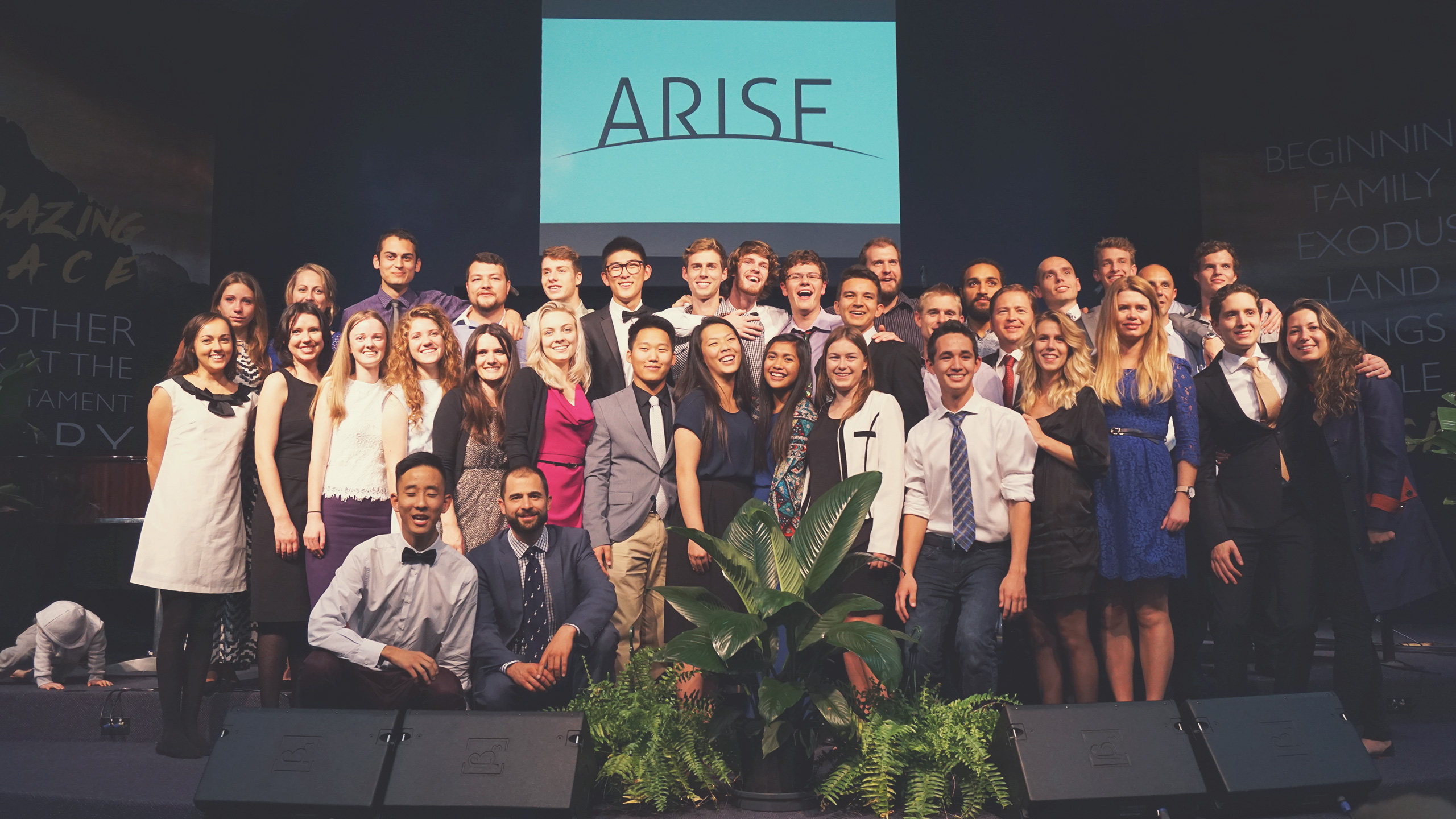 A group of approximately 30 young adults stands on a stage, smiling and posing for a photo in formal and semi-formal attire. Behind them is a screen displaying the word "ARISE." Green plants are at the front of the stage, and dark curtains drape in the background.