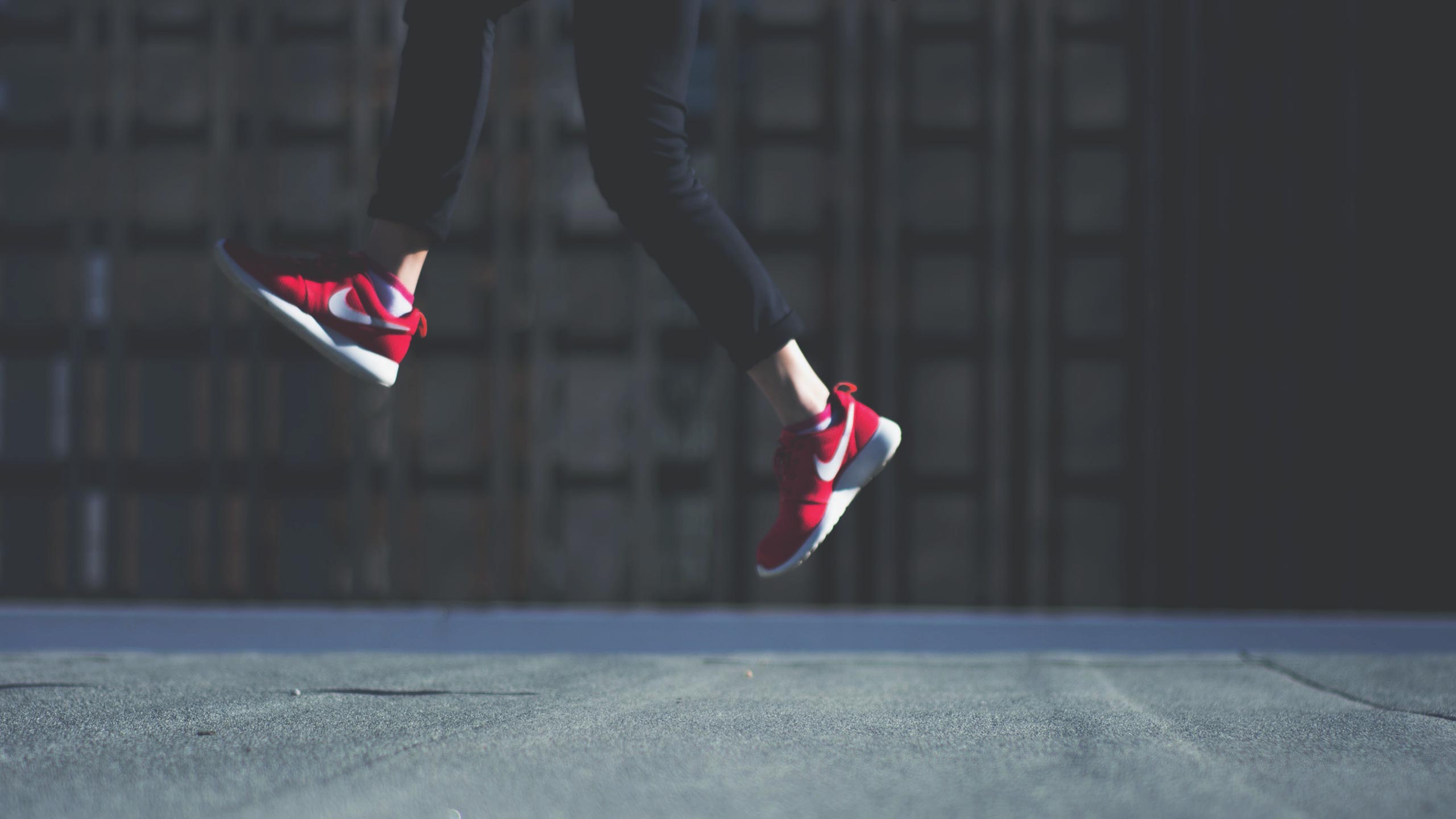 A person is captured mid-air while jumping, wearing red athletic shoes with white accents and black pants. The background is a blurred, dark, outdoor setting.