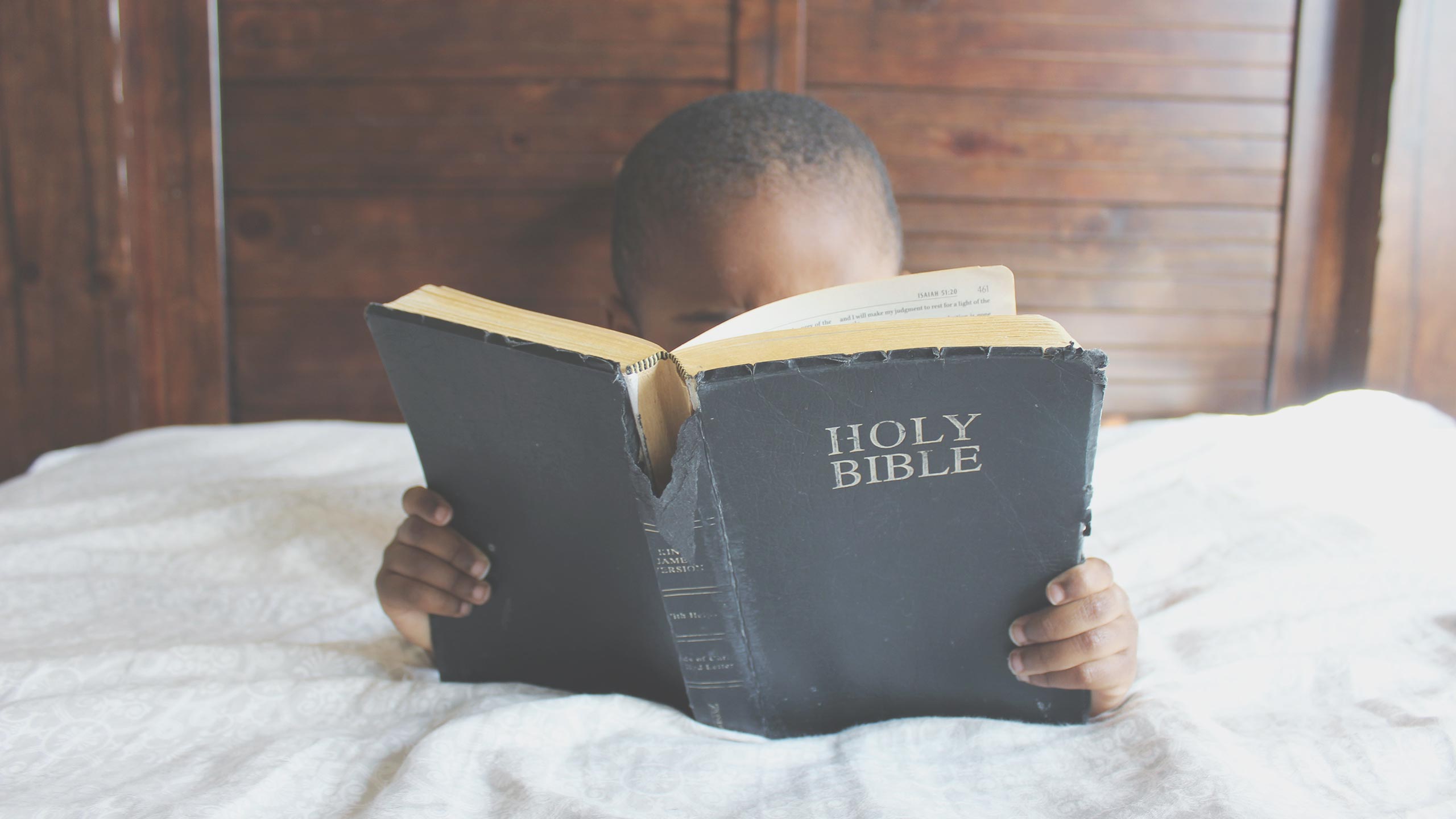 A child sits on a bed, holding and reading a large, worn copy of the Holy Bible. The child's face is partially obscured by the book, with only the top of their head visible. The background consists of wooden panels.