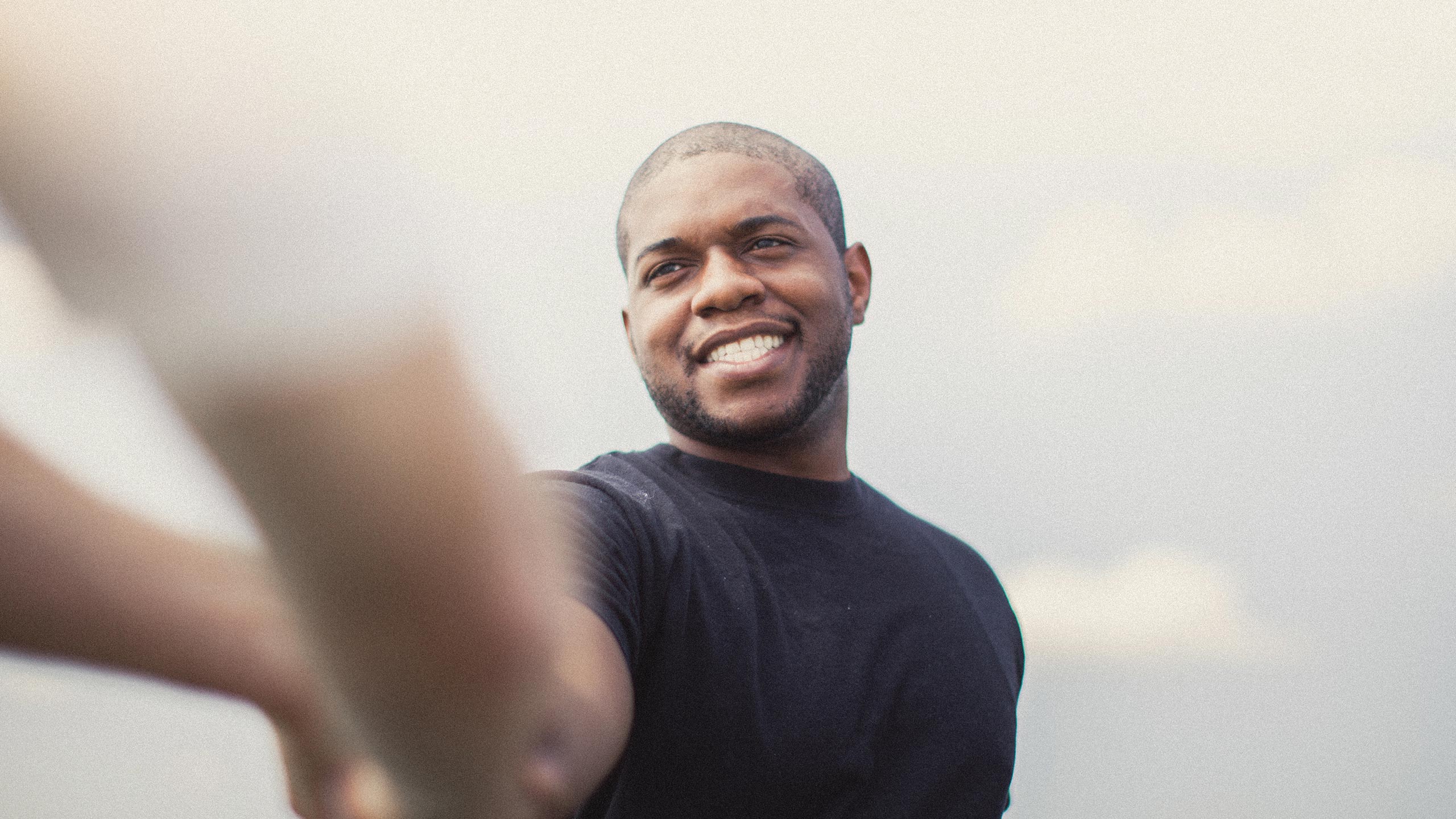 A man with a shaved head and beard smiles warmly while extending his hand forward. He is wearing a black t-shirt, and the background features a pale sky with soft clouds.