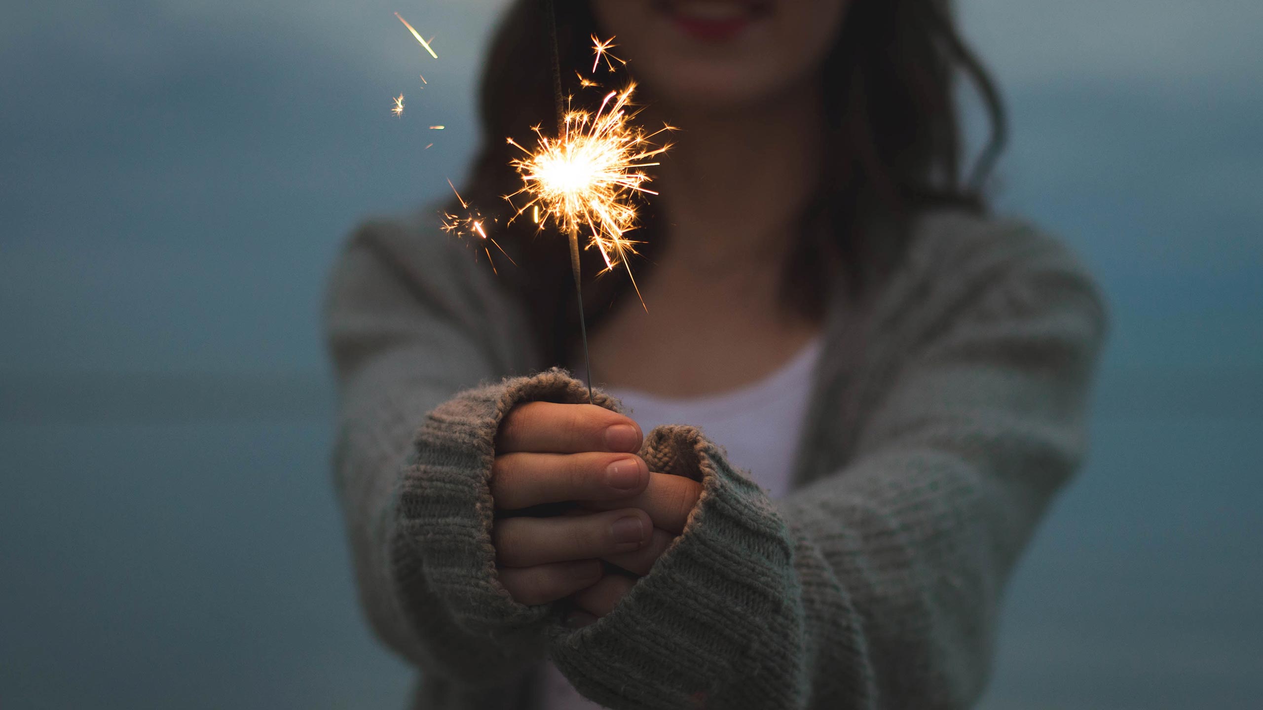 A person with long hair, wearing a gray sweater over a white shirt, holds a lit sparkler. The background is blurred, focusing attention on the sparkler’s bright light and sparks. The scene appears to be outdoors during twilight or evening.