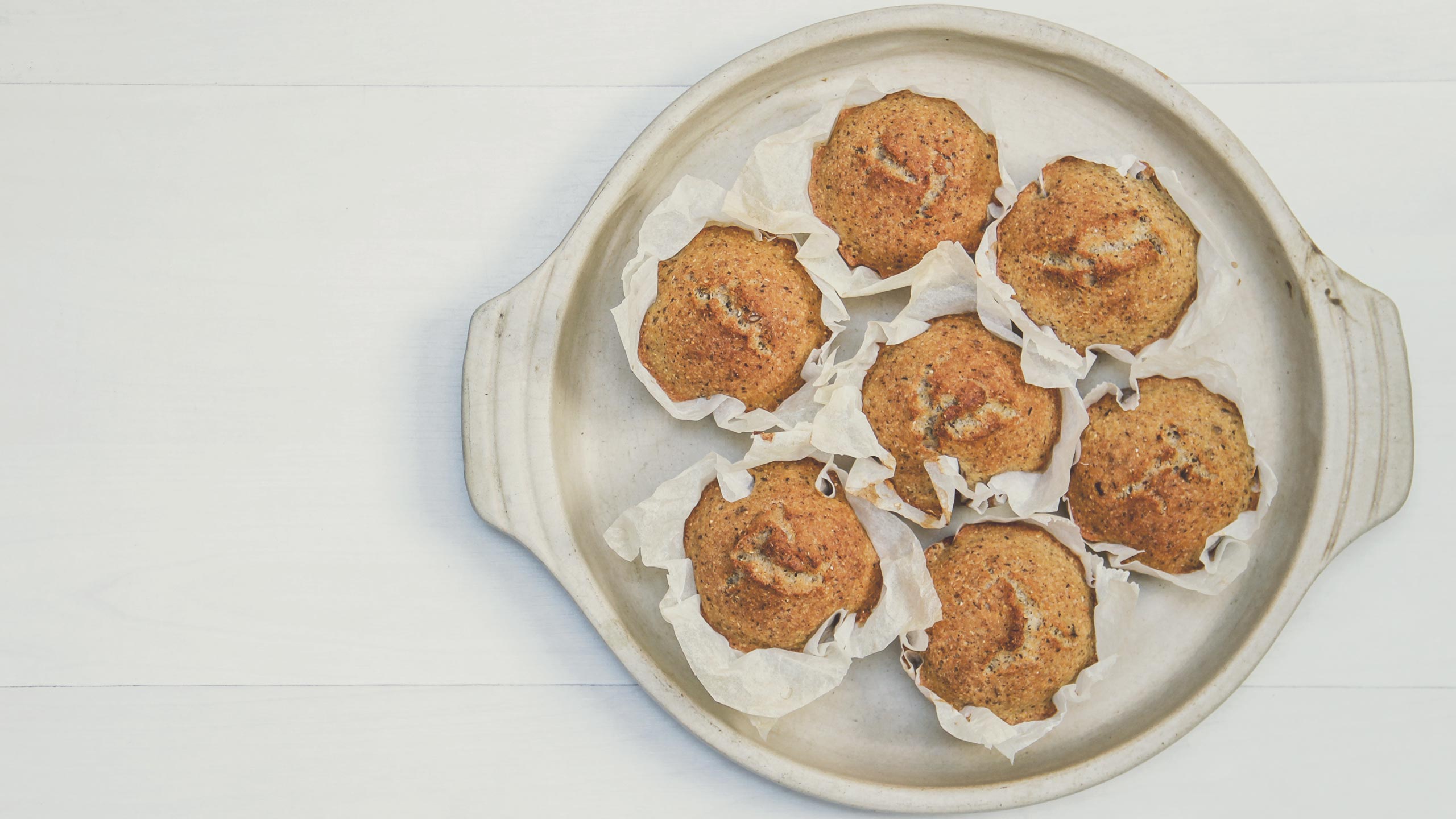 A round ceramic baking dish holds six freshly baked muffins, each lined with white parchment paper. The muffins are golden brown with a slightly textured surface, suggesting they might be topped with nuts or seeds. The dish is placed on a light-colored surface.