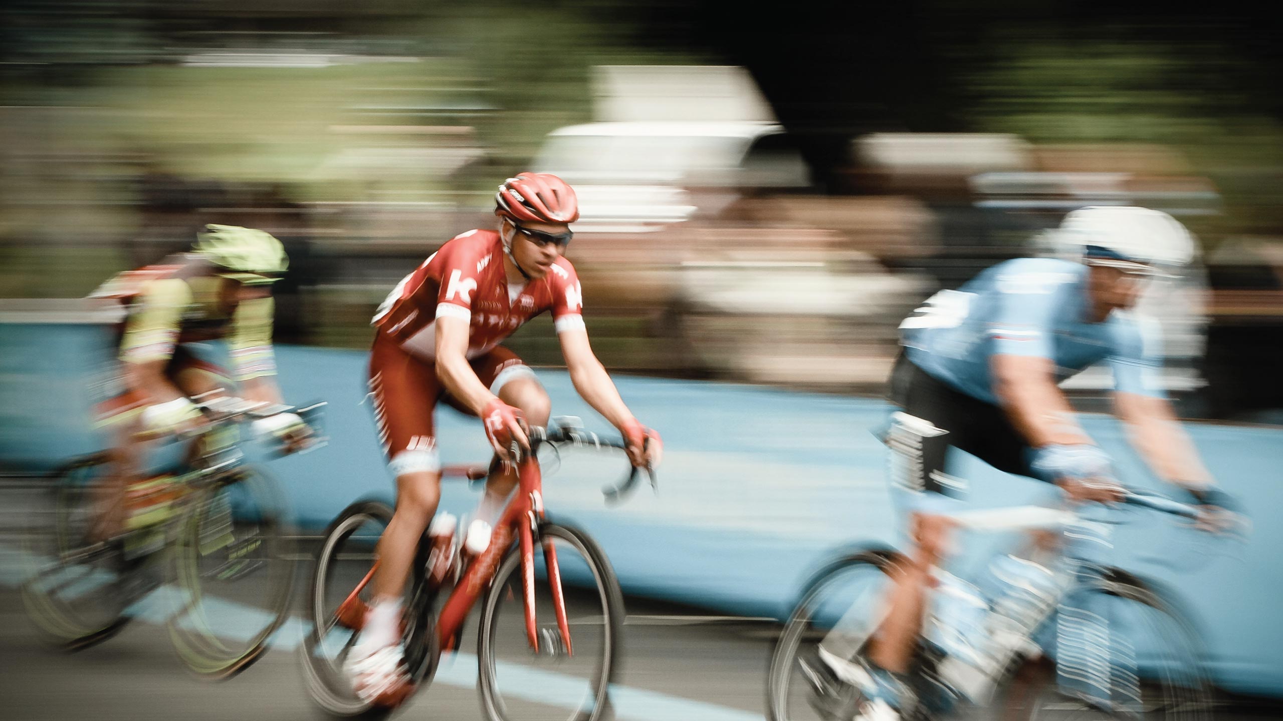Three cyclists, wearing helmets and colorful racing attire, speed down a road during a race. The background is slightly blurred, emphasizing their rapid motion and focus on competing.