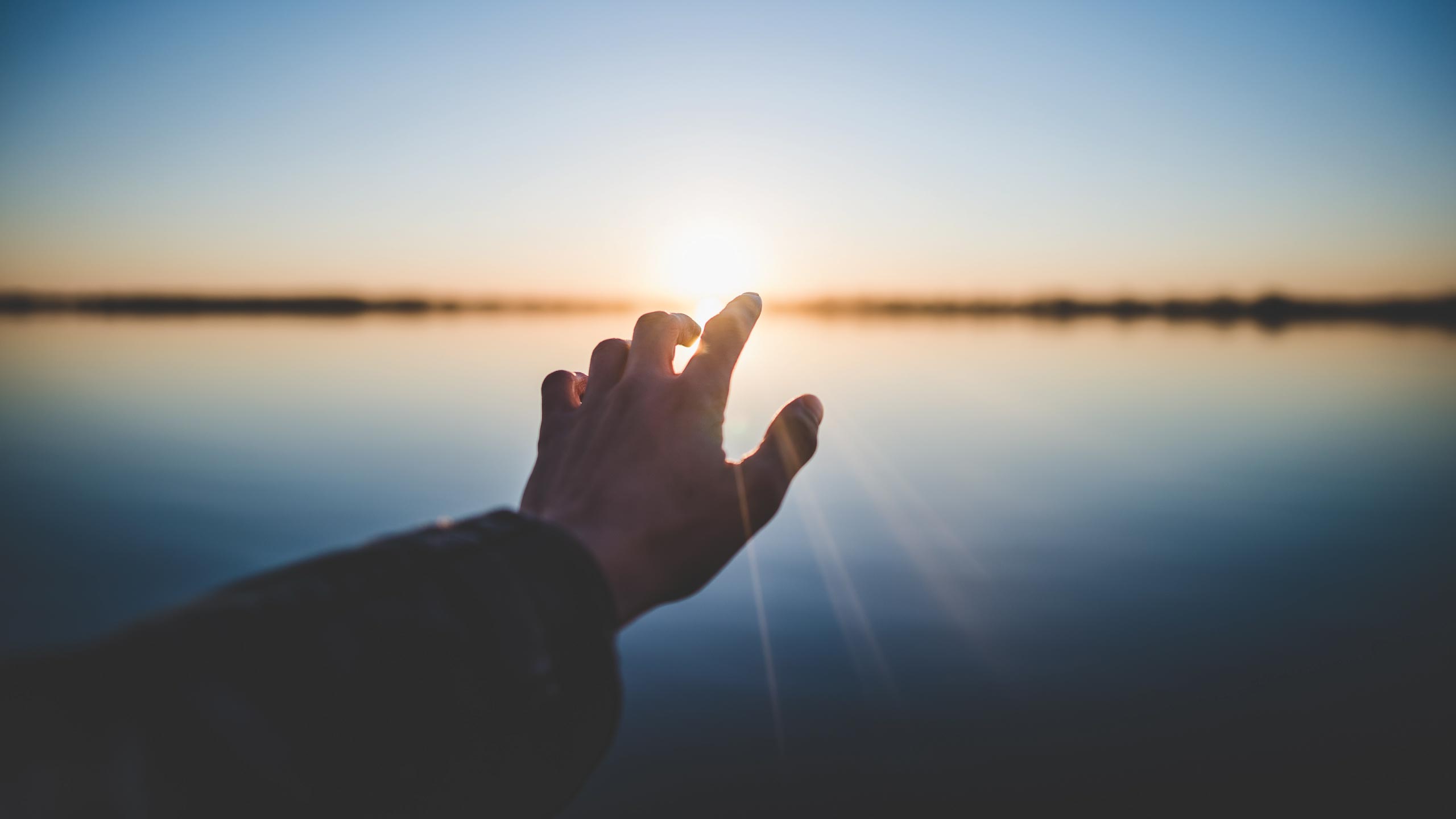 A hand reaches out toward a setting or rising sun over a calm body of water, with the sun's rays creating a serene reflection on the surface. The sky transitions from blue to soft orange hints, evoking a peaceful and contemplative mood.