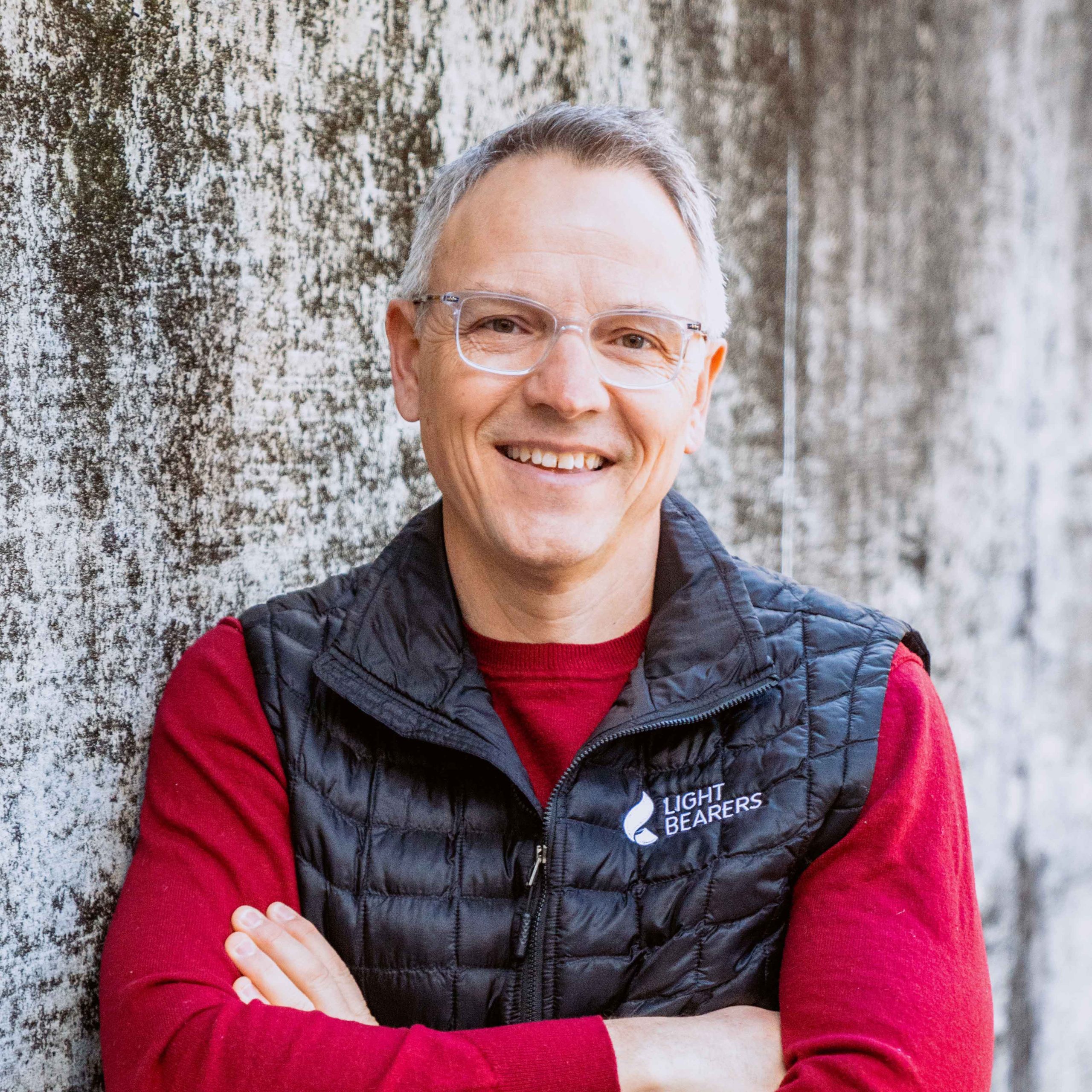 A person wearing glasses, a red sweater, and a black quilted vest with the "Light Bearers" logo stands against a textured wall with arms crossed, smiling at the camera.