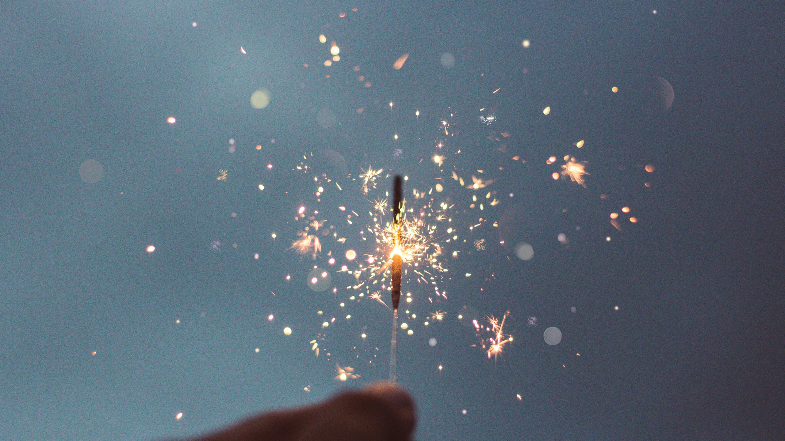 A person holding a lit sparkler against a blurred, soft blue background. The sparkler is emitting bright, colorful sparks and small bursts of light, creating a festive and celebratory atmosphere. The hand holding the sparkler is slightly out of focus.