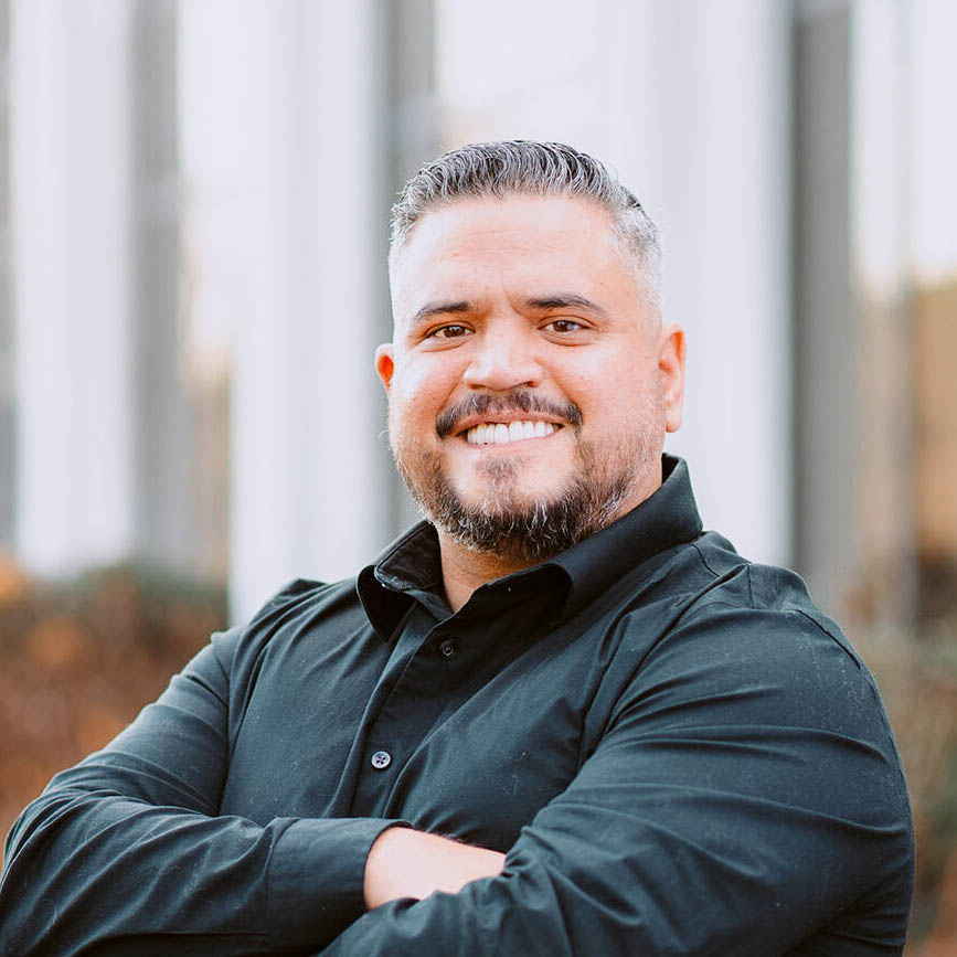 A person with short hair and a beard smiles at the camera with arms crossed, wearing a black long-sleeve shirt. The background is blurred, featuring vertical white structures and an outdoor setting.