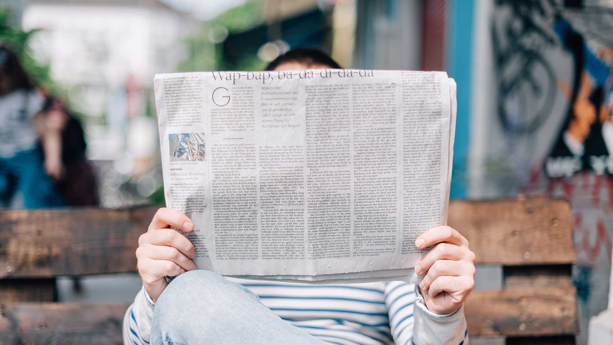 A person sits on a bench, holding and reading a newspaper that covers their face. They are dressed in a striped shirt, and the background shows out-of-focus urban surroundings with hints of graffiti and other people in the distance.