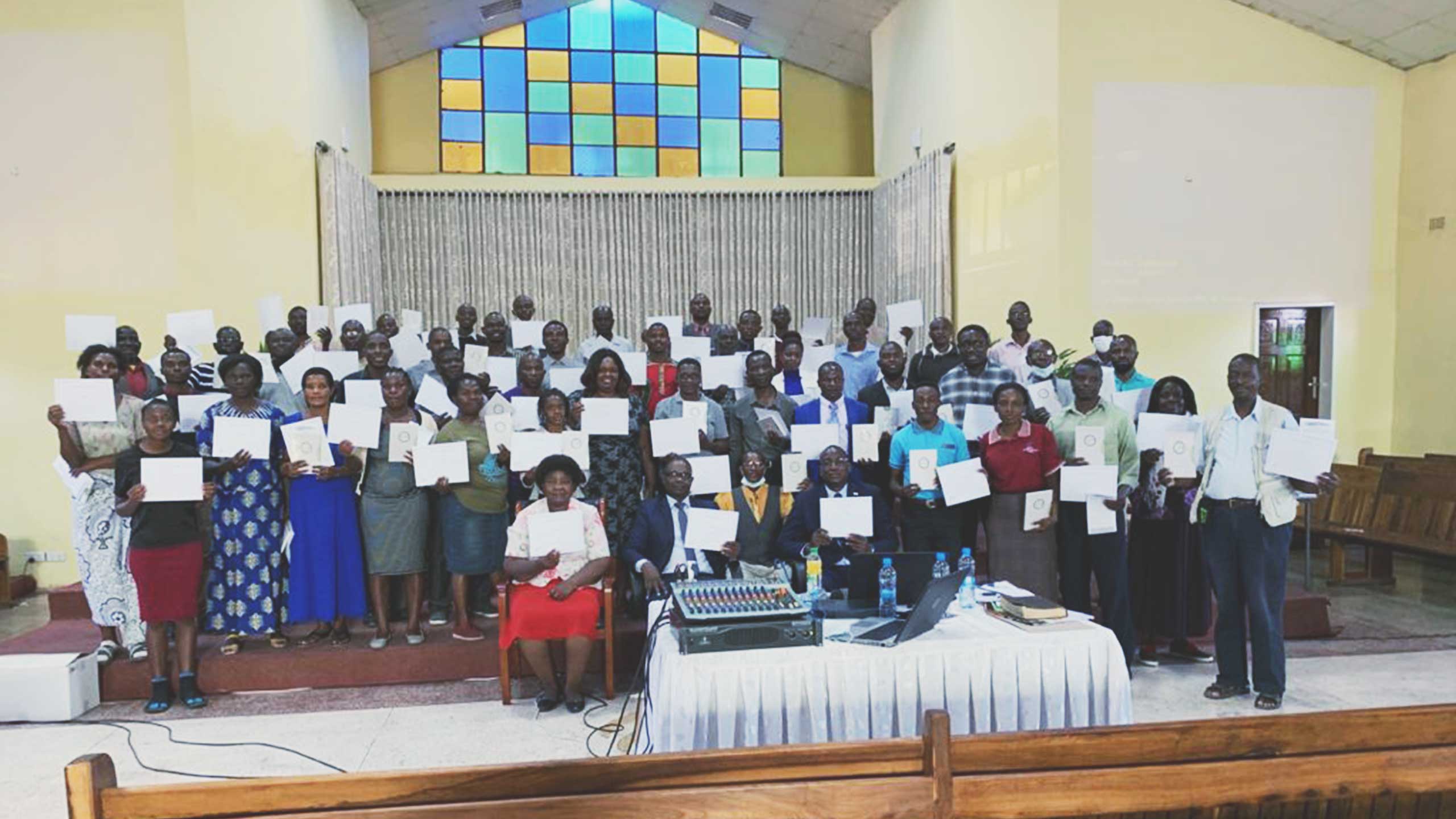 A large group of people poses for a photo inside a building, holding certificates. Behind them is a colorful stained glass window. In front, there is a table with electronic equipment and a few seated individuals. Some wooden benches are visible in the foreground.