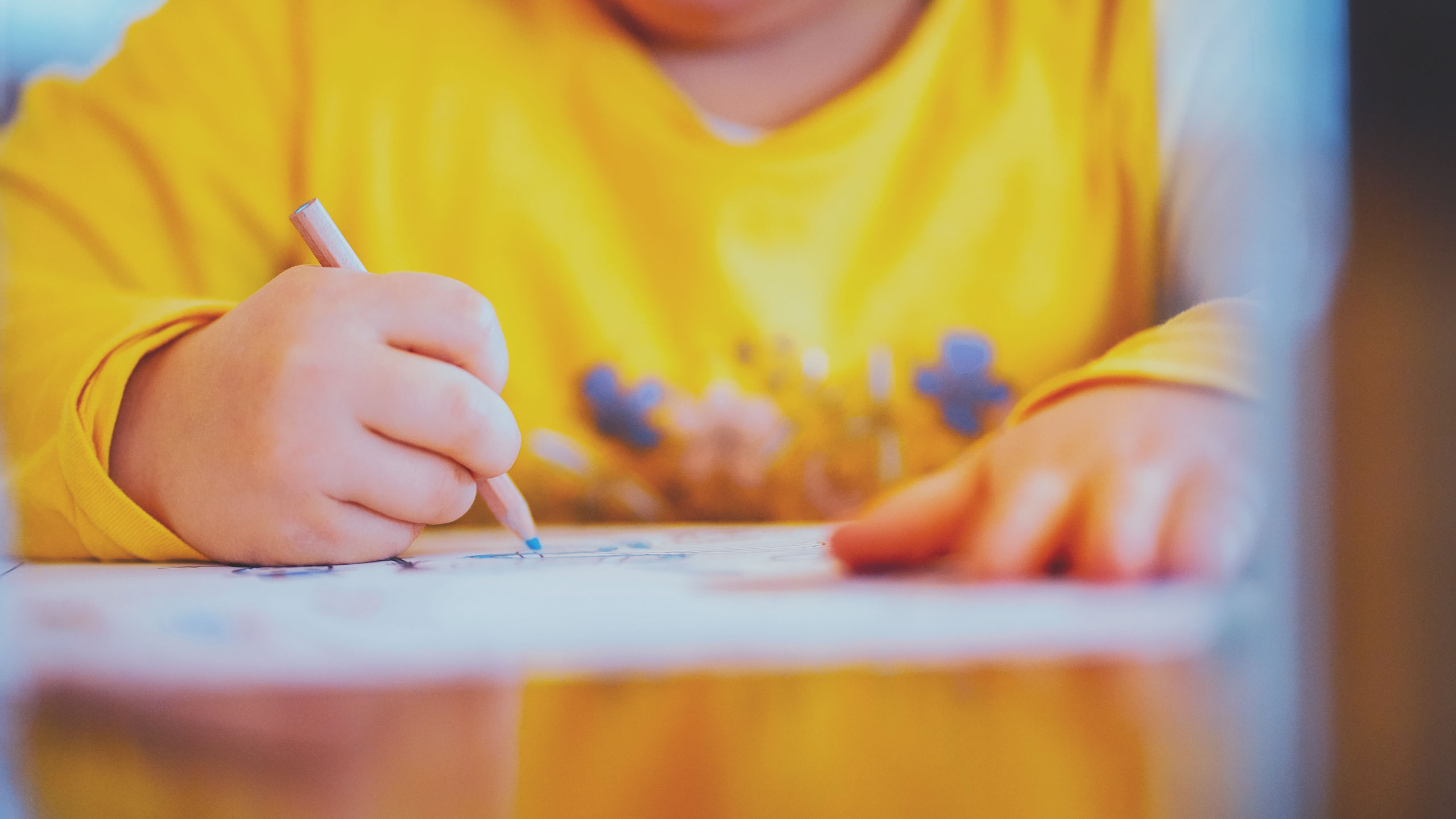 A child in a yellow shirt draws with a pencil on paper. The image focuses on the hands, with the right hand holding the pencil and the left hand resting on the paper. The background is blurry and out of focus.