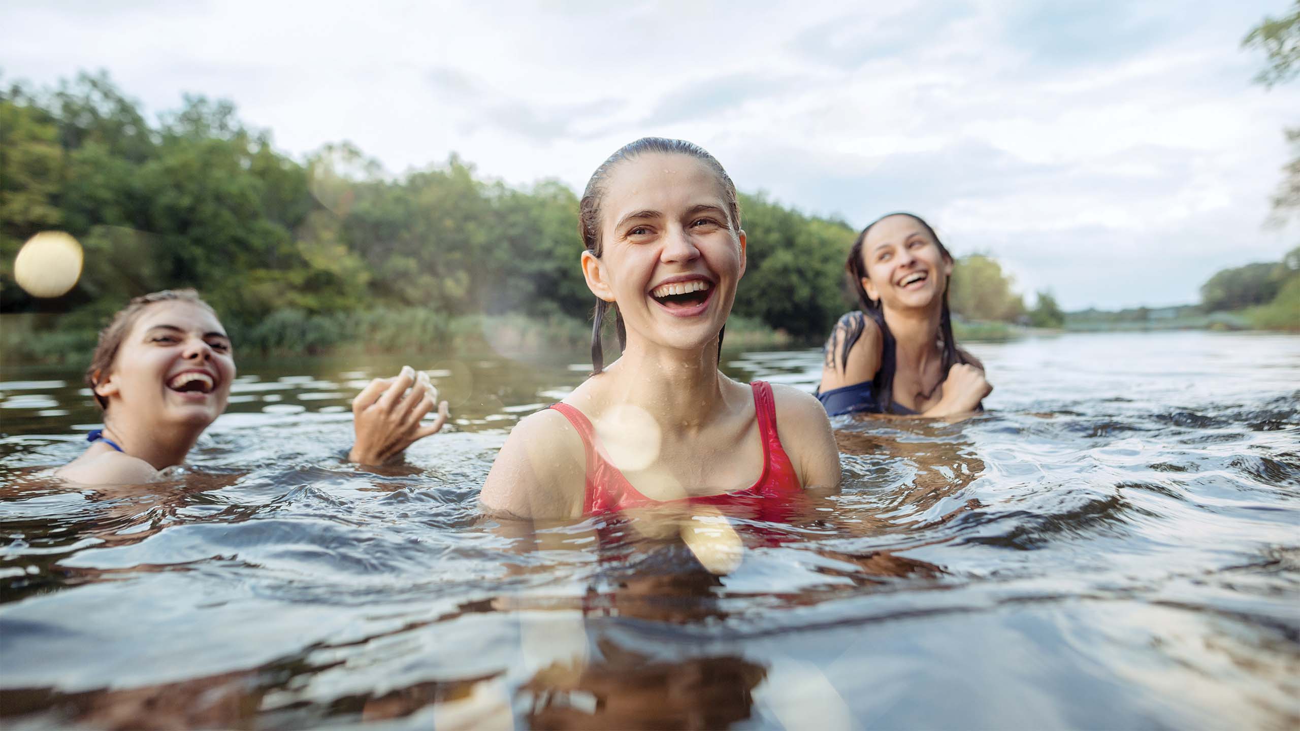 Three people are laughing and enjoying themselves while swimming in a serene body of water surrounded by lush green trees. The person in the center, wearing a red swimsuit, is the focal point, while the other two smile and swim nearby. The sky is partially cloudy.