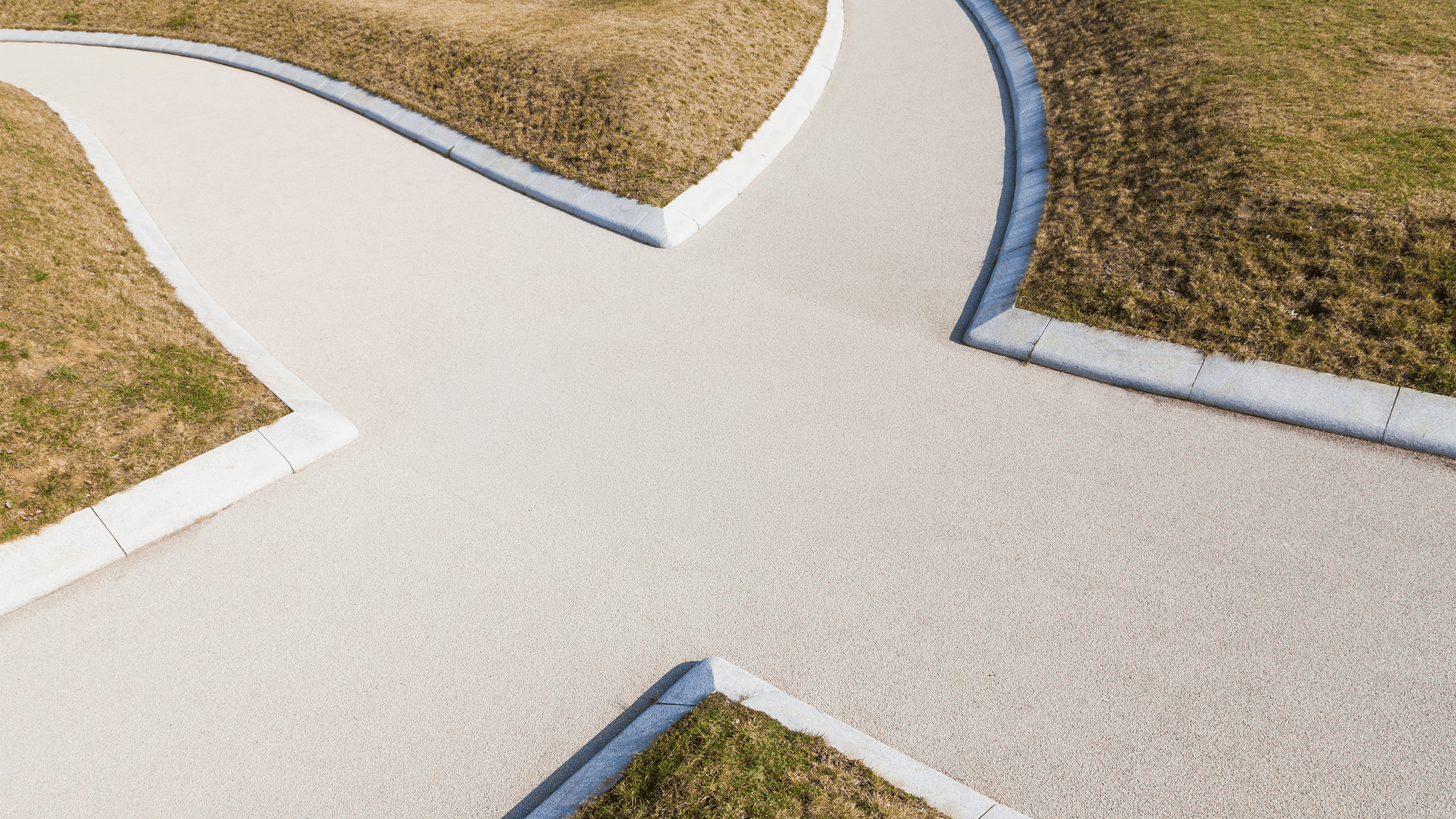 An overhead view of a path splitting into two directions, with neatly trimmed grass and soil on either side, bordered by white curbs. The paths form a 'Y'-shape intersection. The setting appears to be a park or garden area.