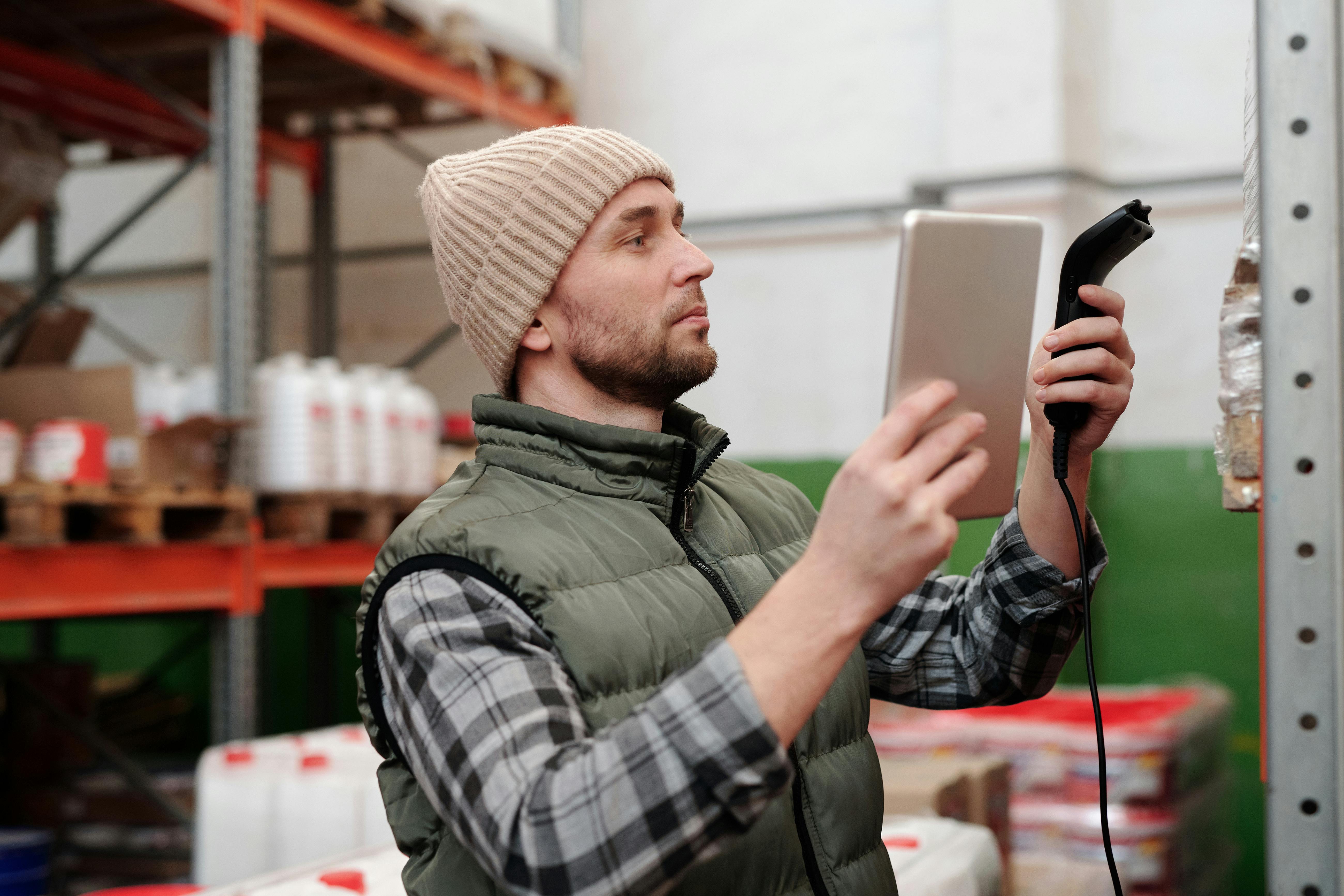 Warehouse worker using handheld scanner and mobile device to conduct inventory count among storage shelves and boxes
