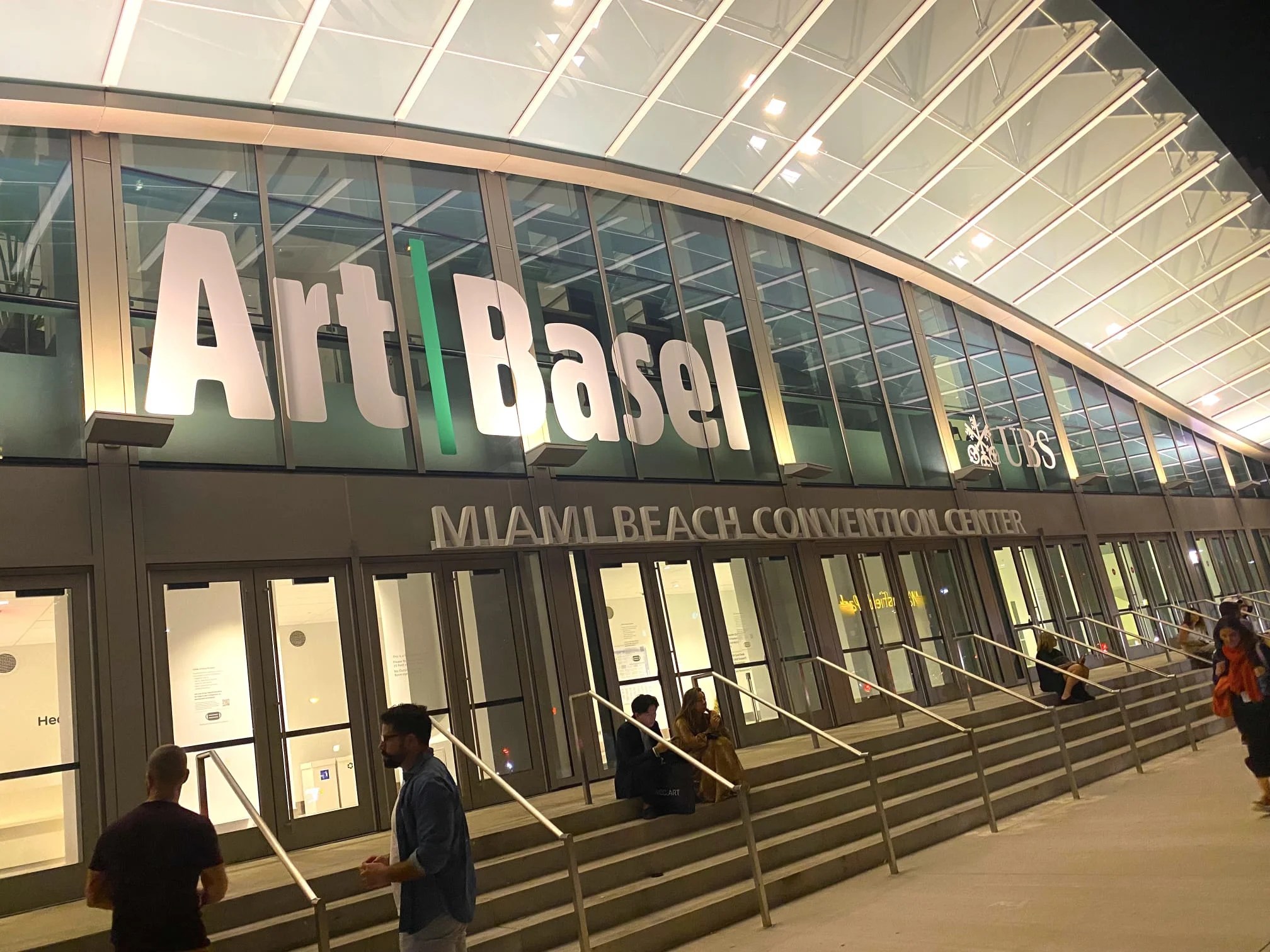 The illuminated Art Basel sign on the curved glass facade of the Miami Beach Convention Center at night, with visitors on the entrance steps.
