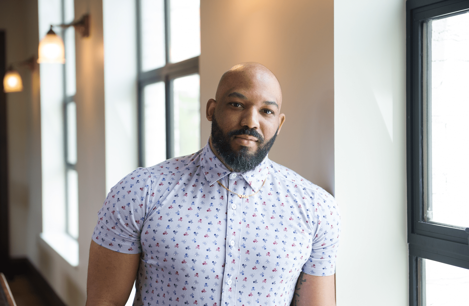 AJ is a bald Black man with a beard leaning against a wall by a window. He is wearing a short-sleeved light blue button-down shirt with small red, white, and dark blue skulls patterned all over the shirt.
