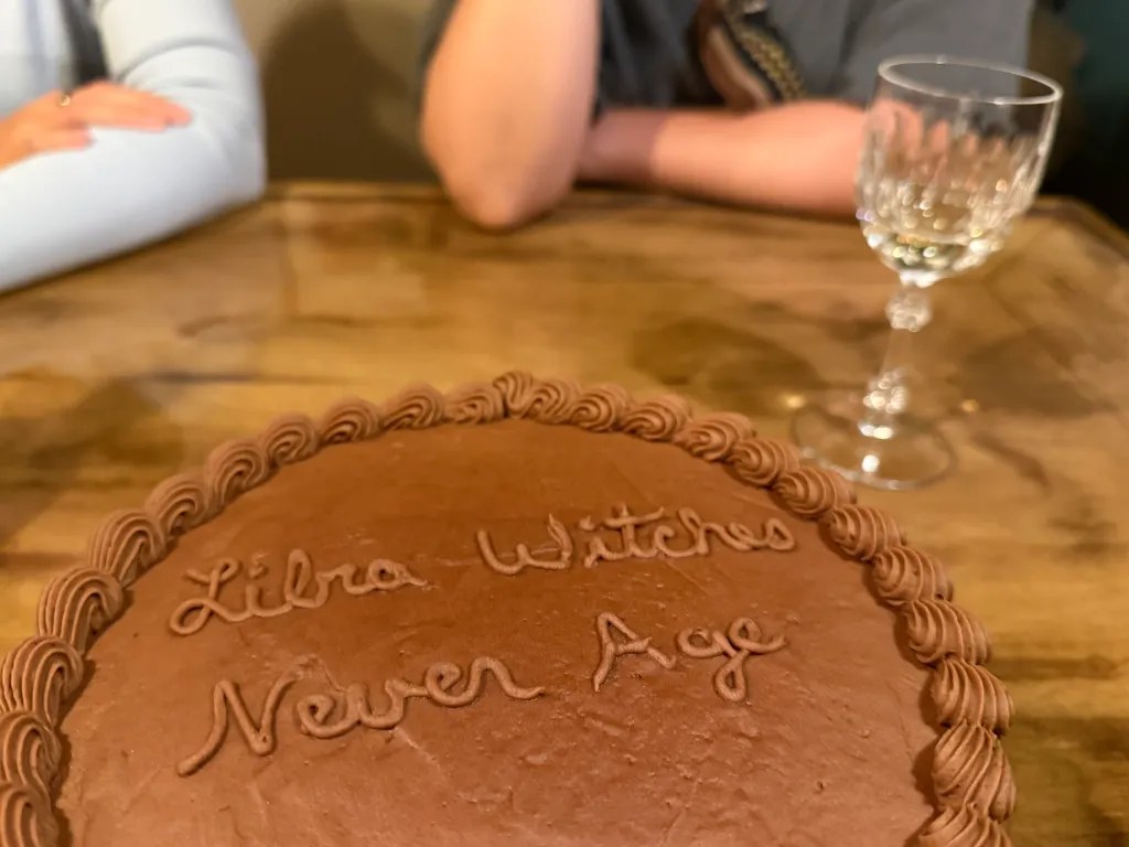 A round chocolate cake with the words 'Libra Witches Never Age' written on top, surrounded by a decorative icing border. In the background, a glass and part of two people's arms are visible.