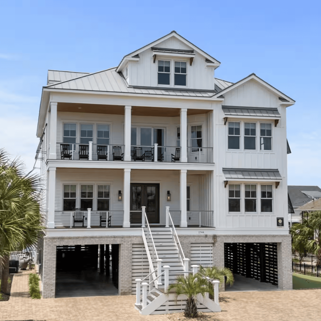 Large coastal-style white beach house on stilts with double-decker covered porches, metal roof, and a staircase leading up to the entrance, surrounded by palm trees.