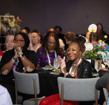Attendees at the World Vitiligo Day-USA conference applaud during a gala dinner. In the foreground, a smiling woman in a red sequined dress and black jacket claps while seated at a table with floral centerpieces, surrounded by other attendees, including several people with visible vitiligo.