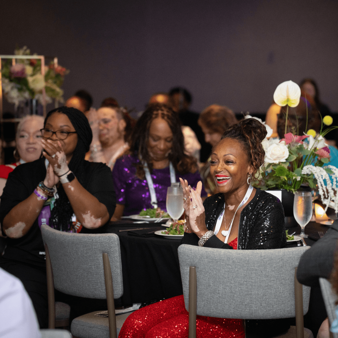Attendees at the World Vitiligo Day-USA conference applaud during a gala dinner. In the foreground, a smiling woman in a red sequined dress and black jacket claps while seated at a table with floral centerpieces, surrounded by other attendees, including several people with visible vitiligo.