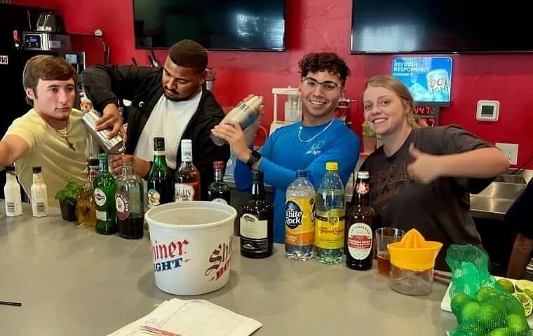 Students practicing cocktail mixing at a bartending school near me, surrounded by various liquors, mixers, and bar tools during a hands-on bartender training session.