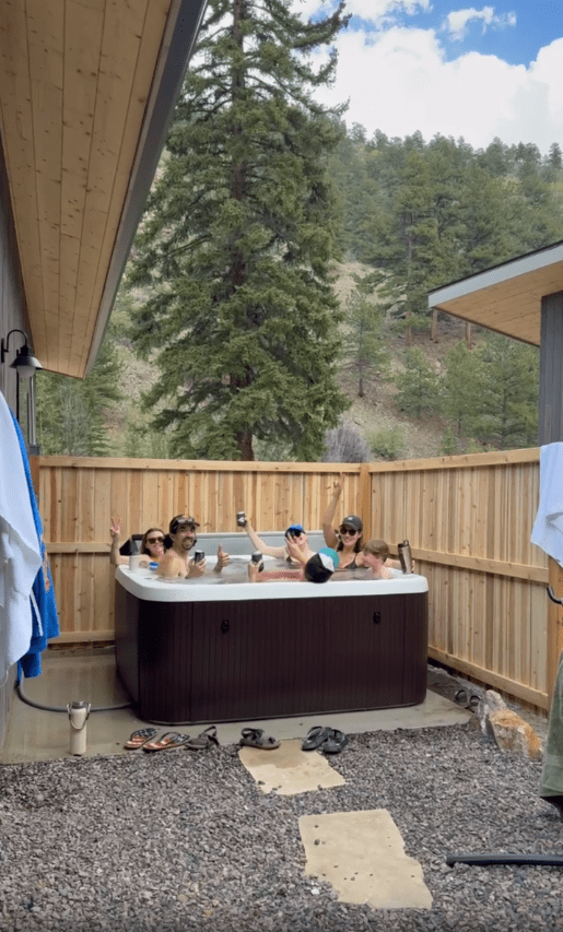 Five people relax and pose for a photo in an outdoor hot tub surrounded by a wooden fence, with trees and hills visible in the background.