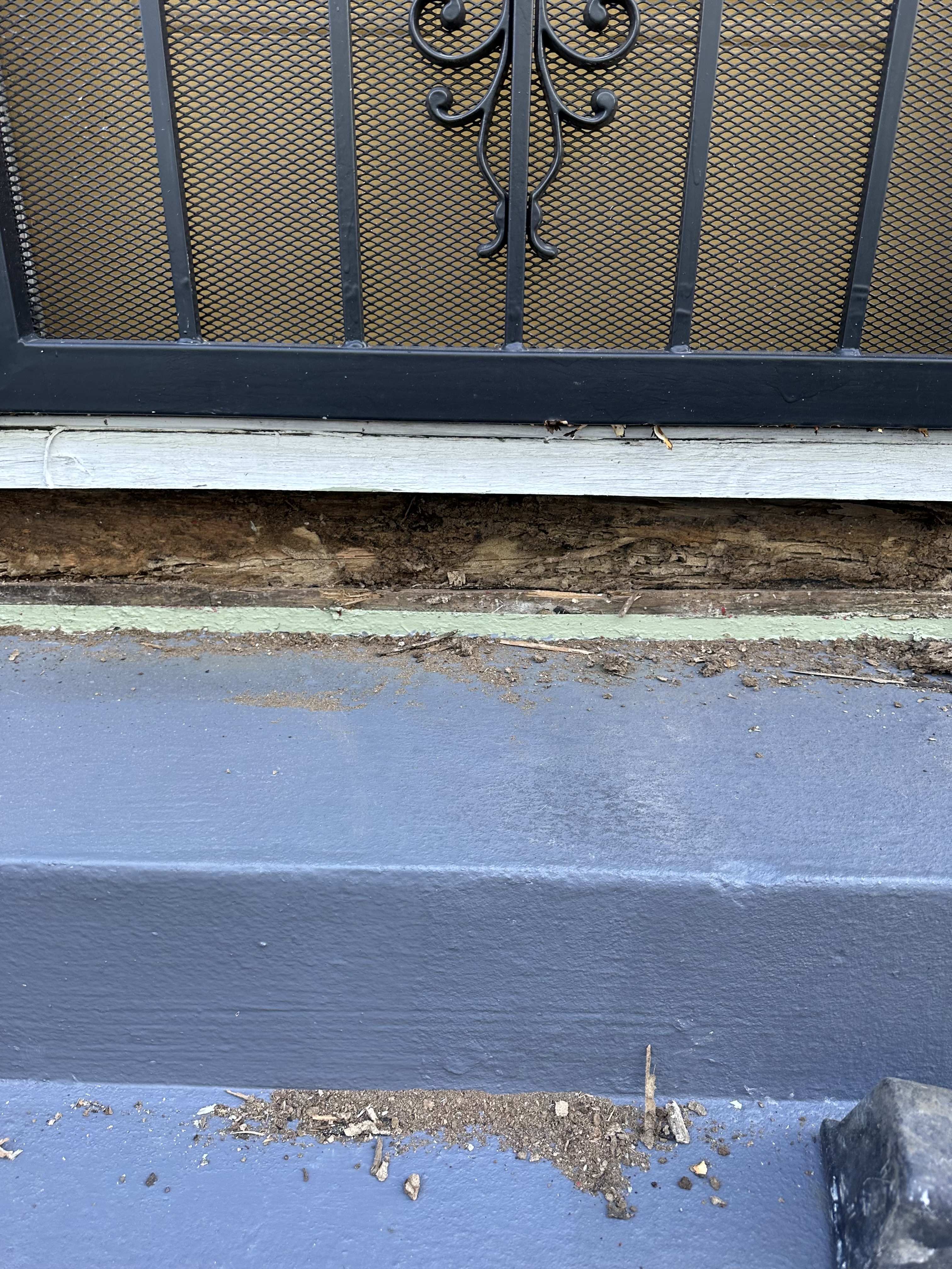 Close-up of a deteriorated windowsill with exposed wood beneath a metal grate. Dust and debris scatter across the blue-painted ledge below.