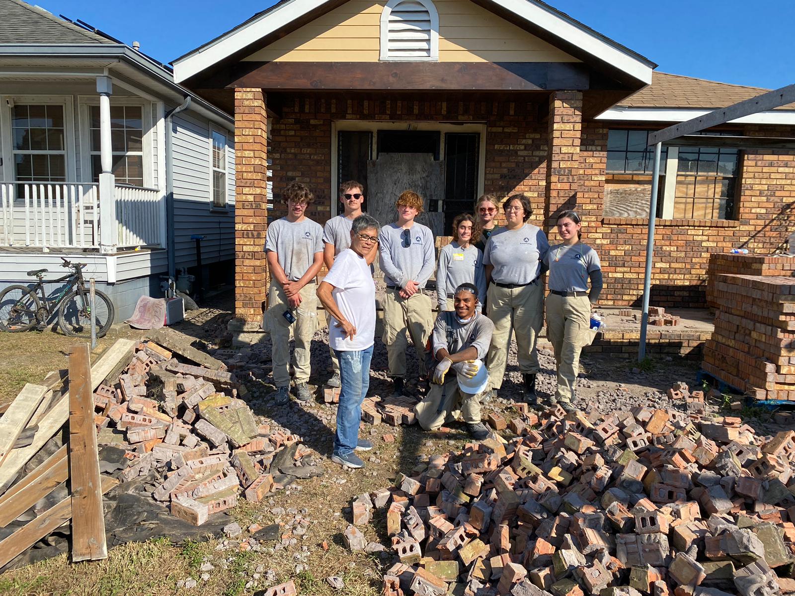 A group of workers stands smiling in front of a brick house under renovation. Piles of bricks are scattered in the yard.