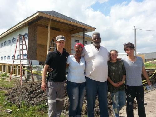A group of five people stands smiling in front of a partially constructed house on a sunny day.