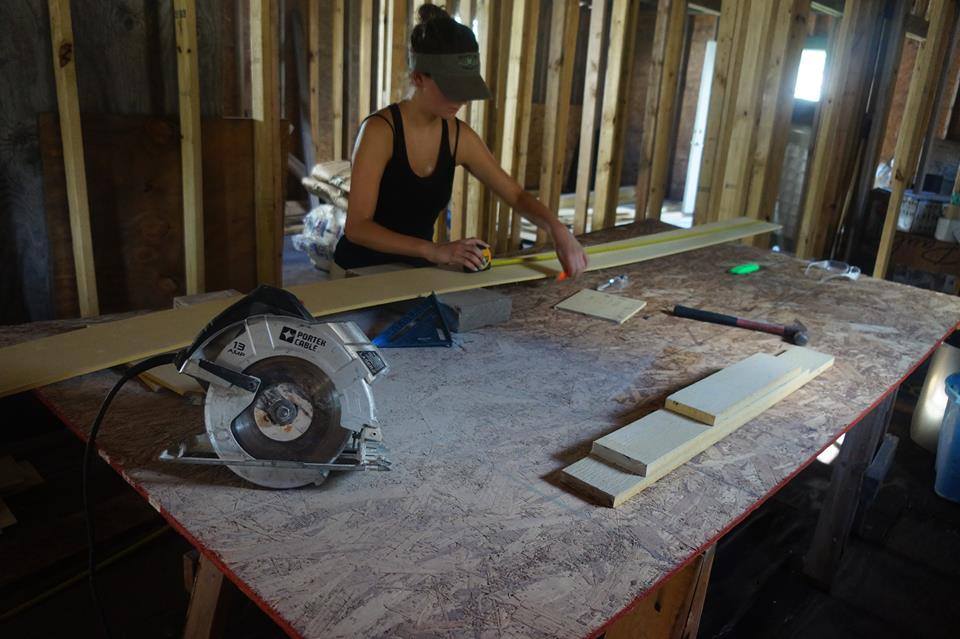 A person in a cap measures wood on a table in a partially built house. A circular saw, hammer, and wood pieces are on the table.