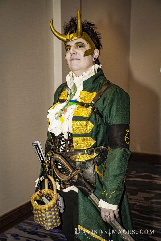 A person in elaborate cosplay attire, featuring a green coat with gold accents, a ruffled white shirt, and horned headgear, posing confidently in a hallway.