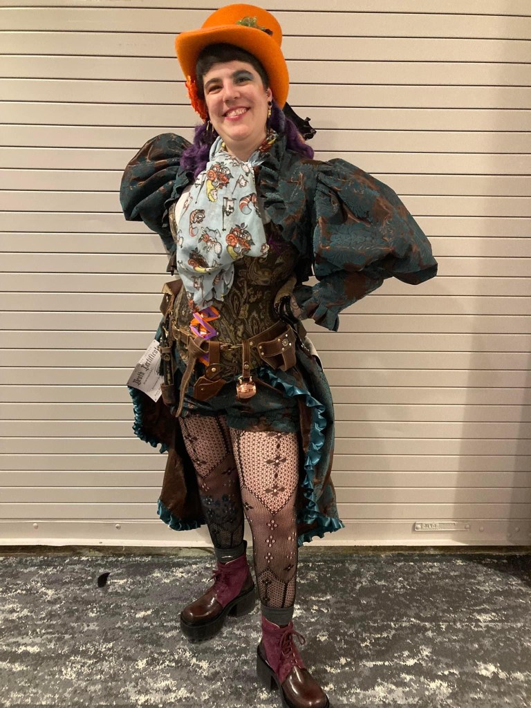 A person dressed in a colorful and elaborate steampunk-inspired outfit, featuring a large orange hat, a ruffled blouse, and patterned leggings. The individual is striking a playful pose in front of a neutral background.
