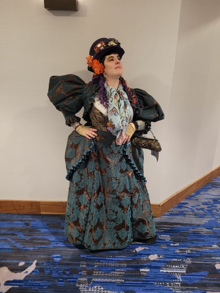 A person in a decorated Victorian-inspired costume with a large, patterned dress, a matching hat, and colorful accessories, striking a pose against a neutral wall.