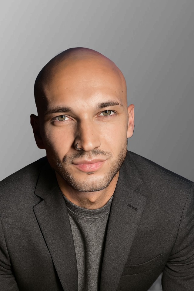 Porträt eines jungen Mannes mit glatze, der einen schwarzen Blazer trägt. Der Mann schaut direkt in die Kamera und hat einen neutralen Gesichtsausdruck. Der Hintergrund ist unscharf und grau.