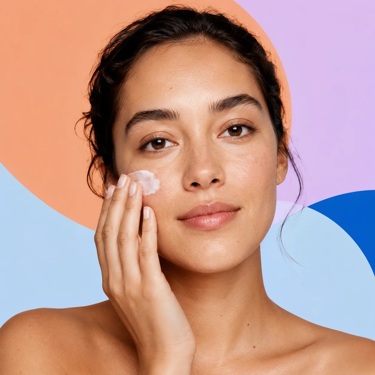 Woman applying moisturizer to her cheek against apricot and blue shapes