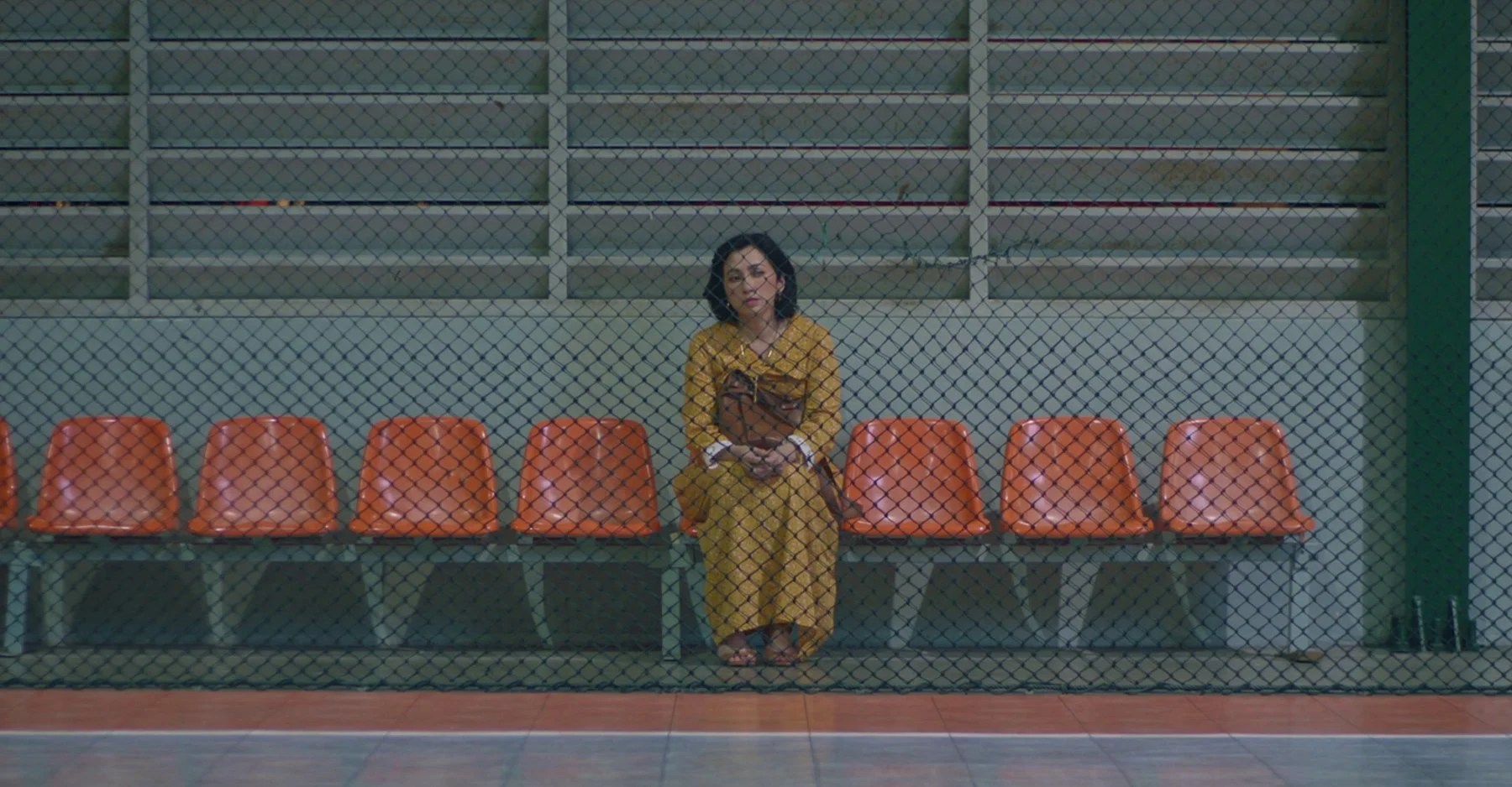 Still from Maryam Pagi ke Malam. A woman in a yellow dress sits alone on a bench in front of a chain-link fence.