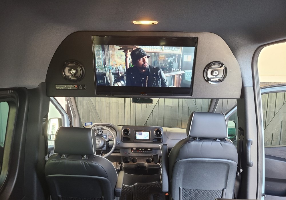 Interior of a luxury van with a ceiling-mounted flat-screen TV and speakers above the driver's cab and leather seats