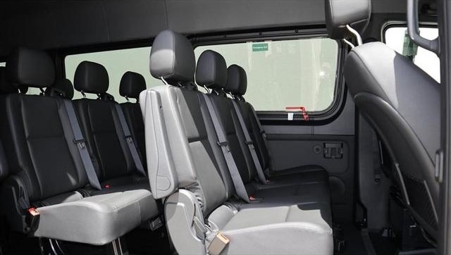 Interior of a passenger van showing multiple rows of gray and black seats with seatbelts and large side windows