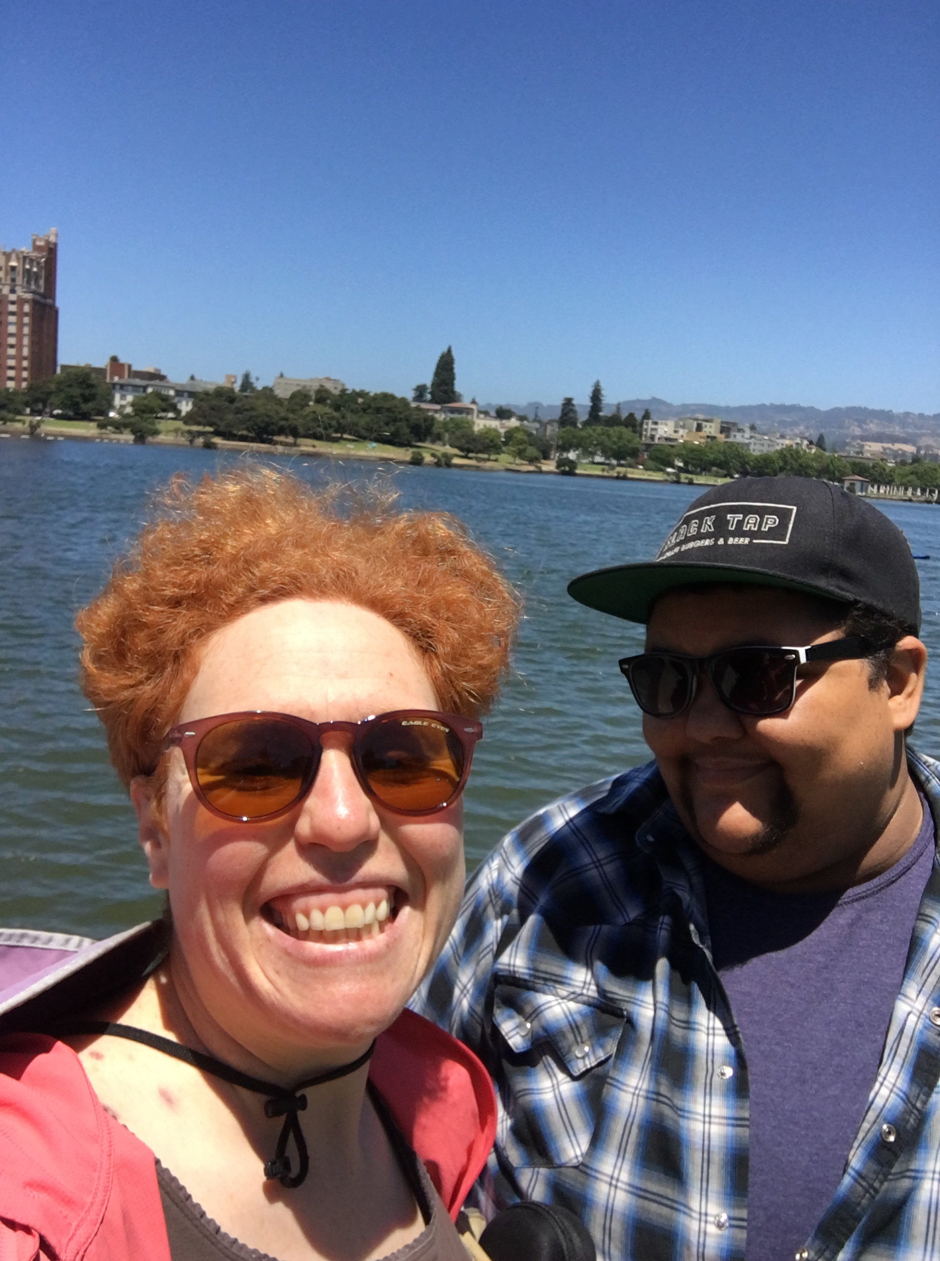 L: Ma'ayan wearing sunglasses & smiling; R: Andy in sunglasses and baseball cap. Water in background. M has redhair and fair skin. A has brown skin and goatee.