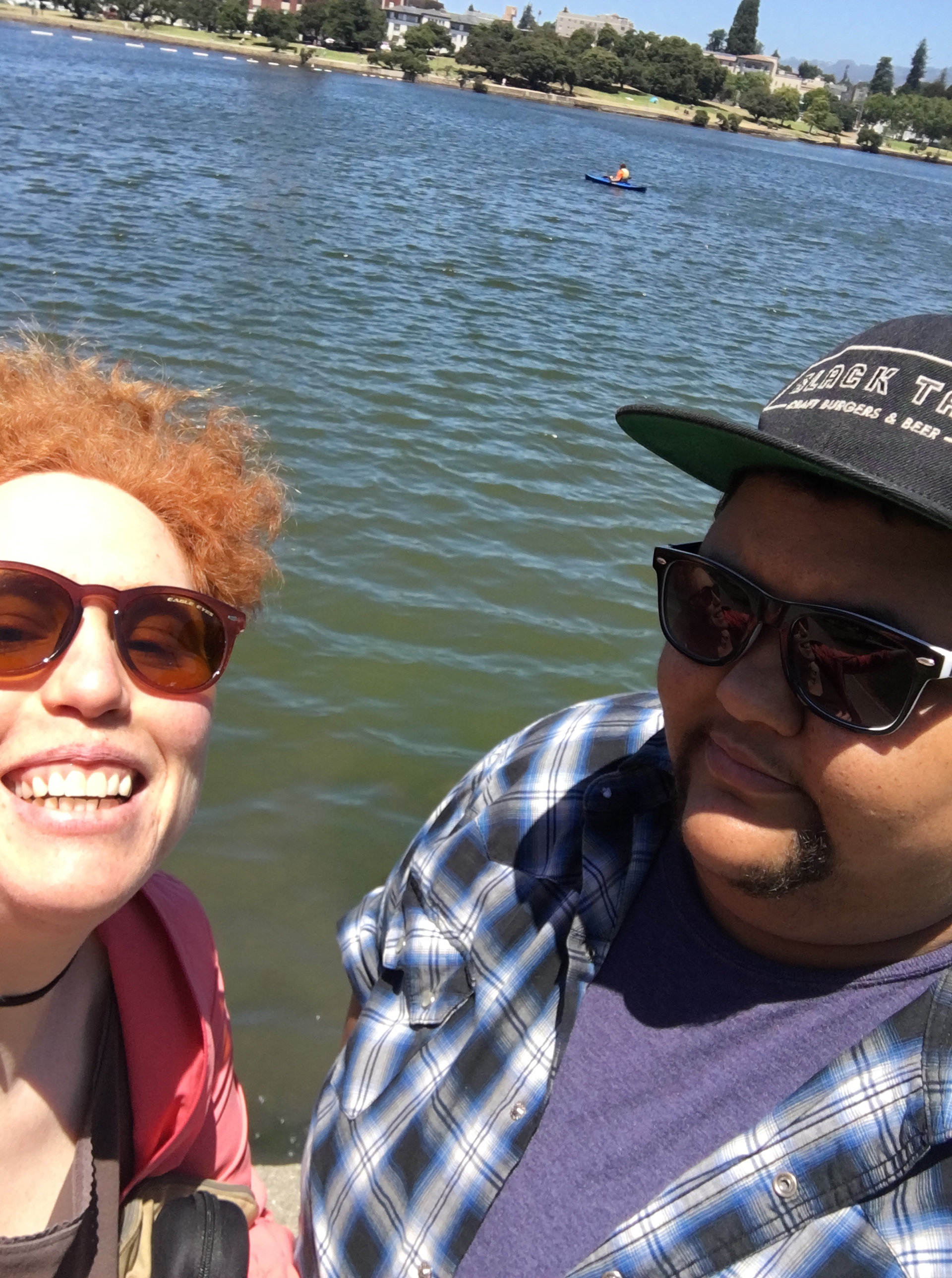 L: Ma'ayan wearing sunglasses & smiling; R: Andy in sunglasses and baseball cap. Water in background. M has redhair and fair skin. A has brown skin and goatee.