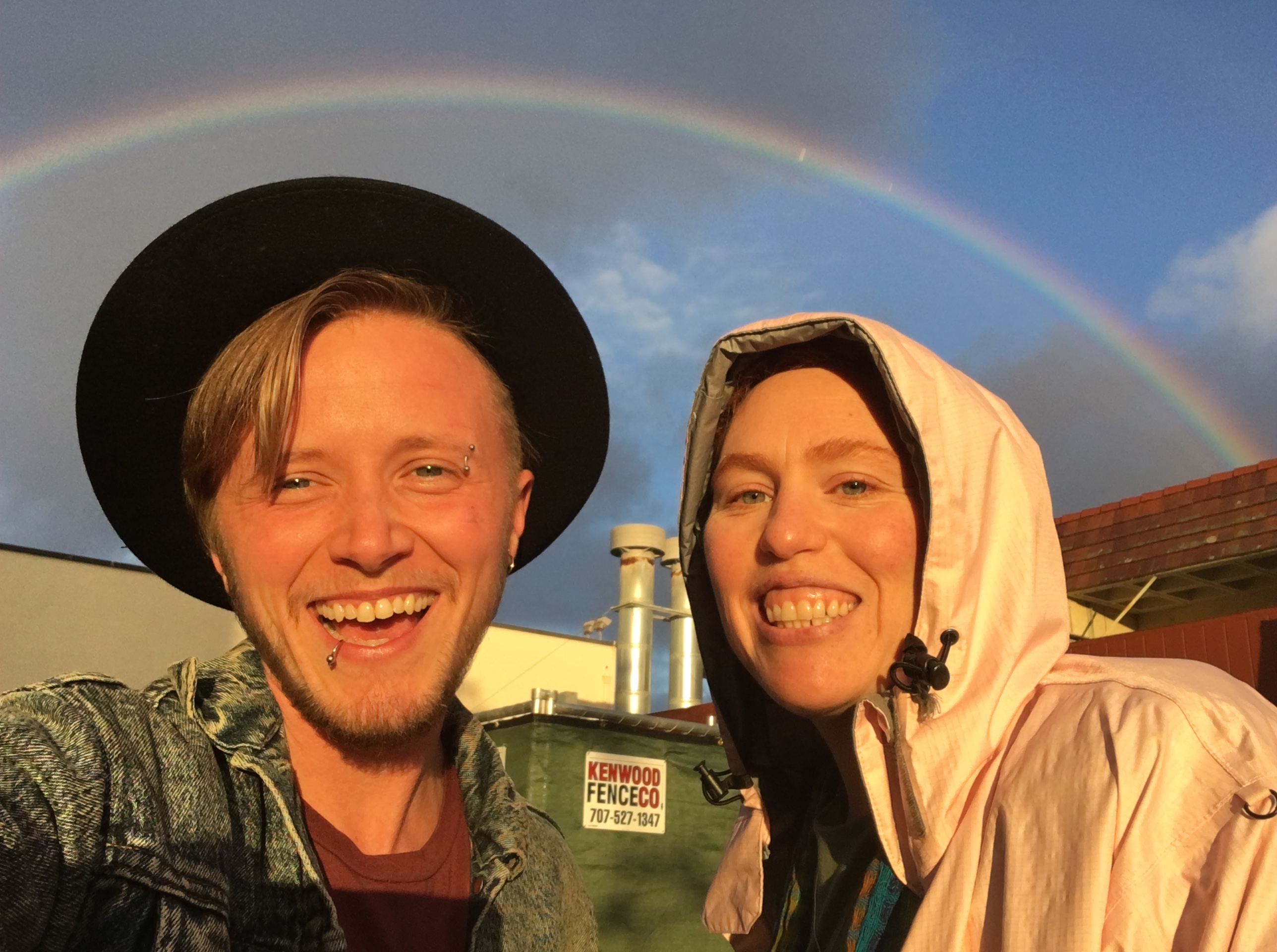 L: Freddie has fair skin, lip ring and is wearing widebrim black hat; R: Ma'ayan has fair skin, smiling and in pink raincoat w/hood over head. Rainbow in backfround!