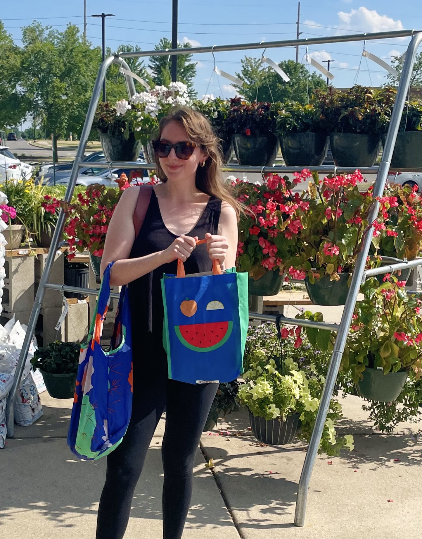 A woman stands outside in front of flower displays. She holds two reusable bags