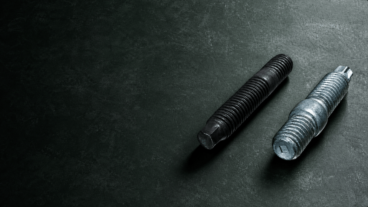 Two large metal bolts with helical threads lying on a dark textured surface.